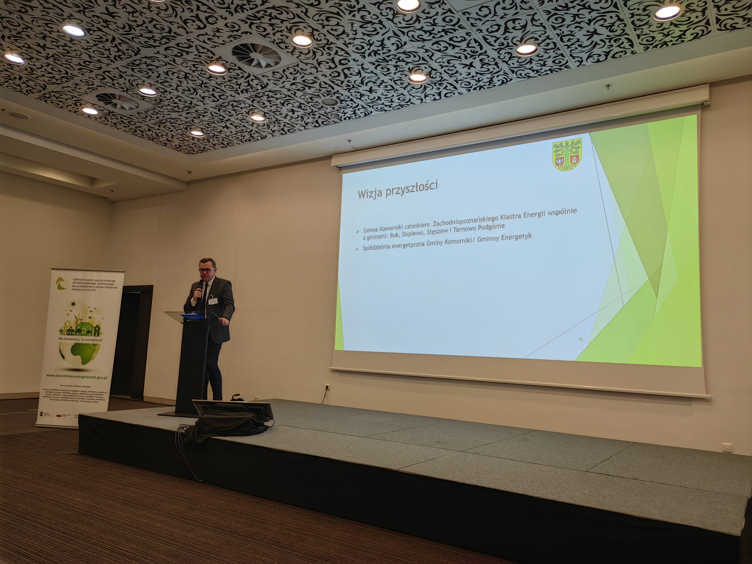 1 osoba stoi na scenie w trakcie konferencji i przemawia do uczestników.