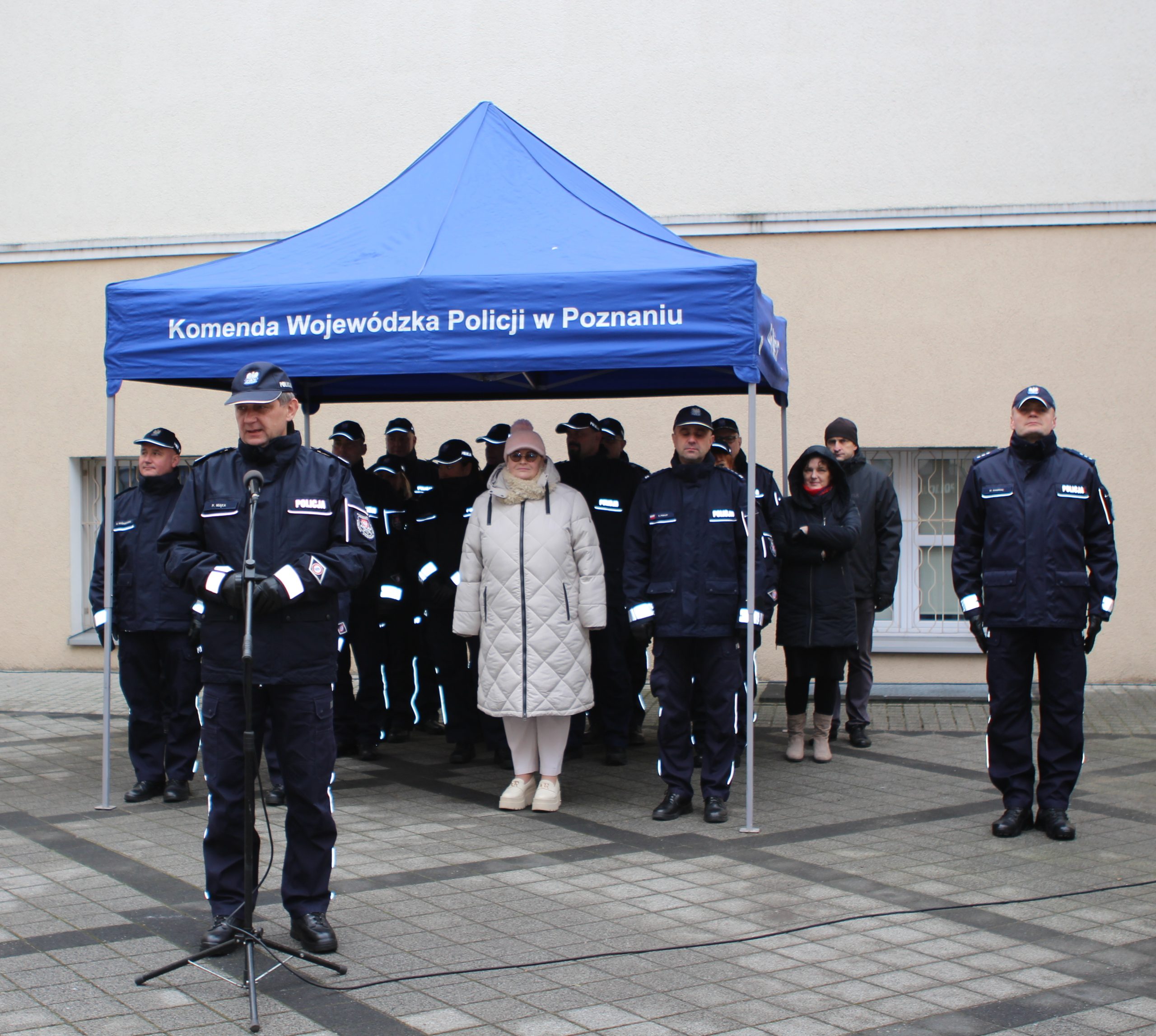 Goście zaproszeni na uroczystość przekazania samochodów bojowych dla Komendy Wojewódzkiej w Poznaniu, stoją pod namiotem na dziedzińcu podczas uroczystości.