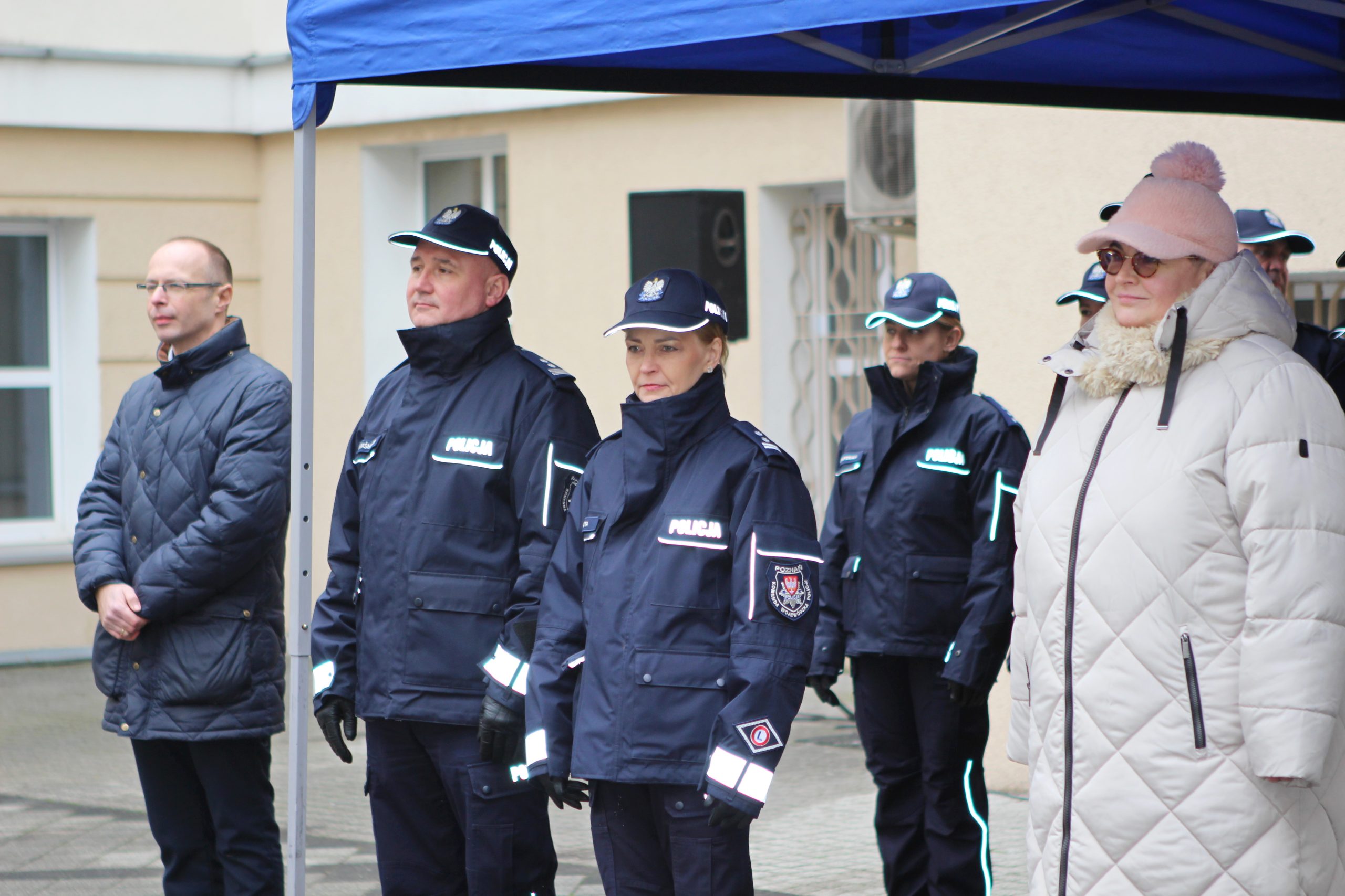Goście zaproszeni na uroczystość przekazania samochodów bojowych dla Komendy Wojewódzkiej w Poznaniu, stoją pod namiotem na dziedzińcu podczas uroczystości.