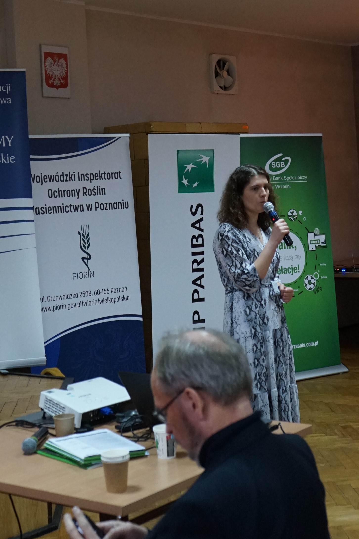 Prelekcja Doradcy Energetycznego podczas Wielkopolskiego Forum Rolniczego - powiat wrzesiński. Jedna osoba stoi na tle bannerów reklamowych, z mikrofonem w dłoni i przemawia.