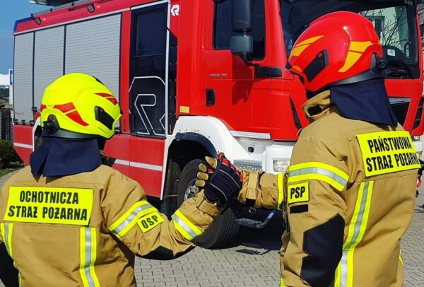Zdjęcie przedstawia dwoje strażaków - jednego z Ochotniczej Straży Pożarnej, a drugiego z Państwowej Straży Pożarnej, stojących na tle wozu strażackiego i podających sobie ręce.