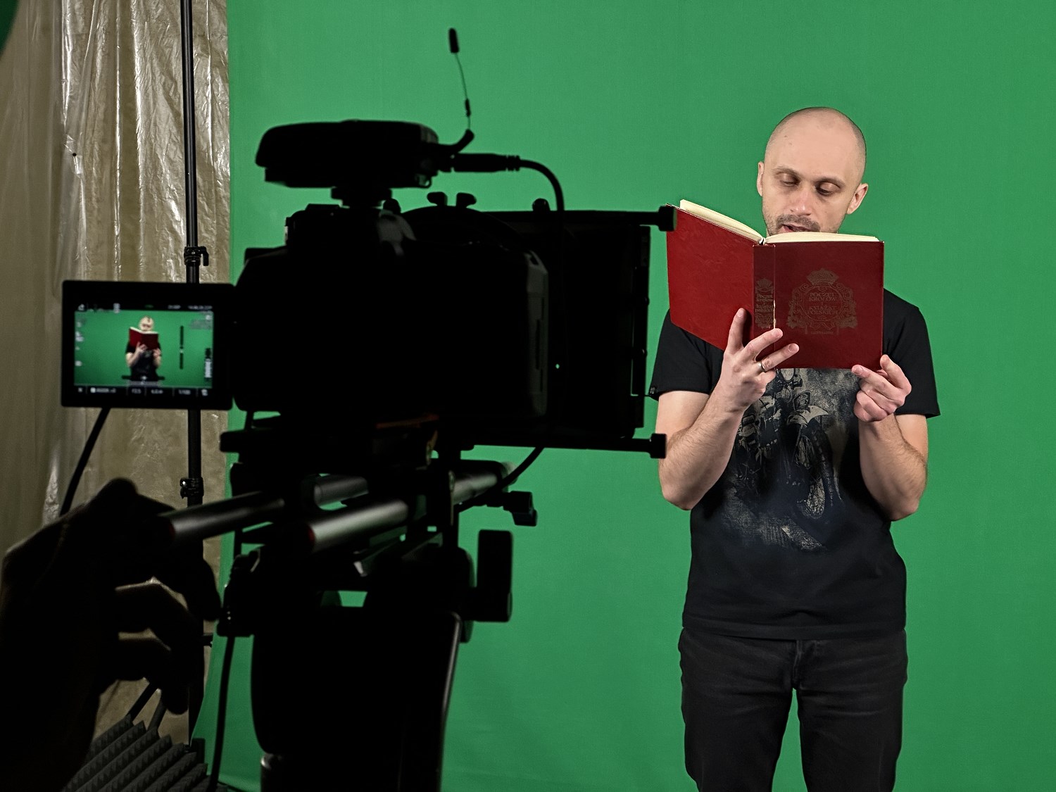 Jedna osoba stoi na tle zielonej ściany i czyta z książki przed kamerą, która nagrywa.
