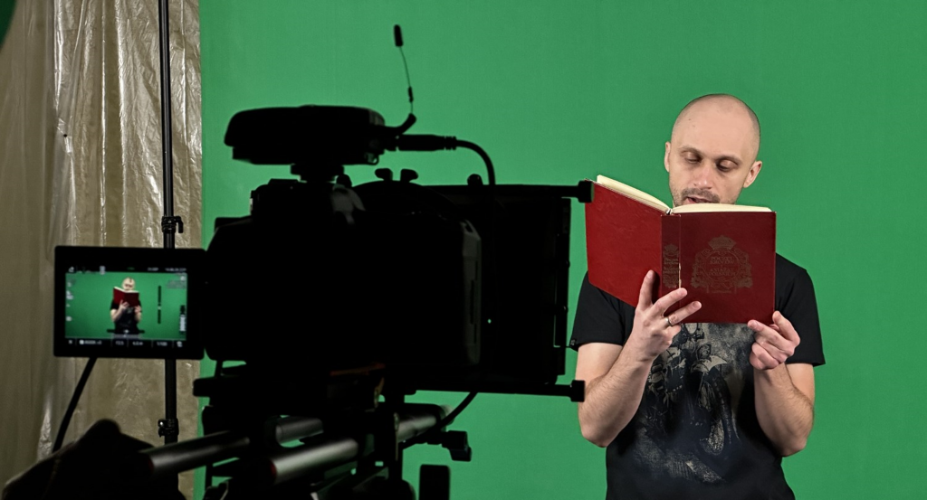 Jedna osoba stoi na tle zielonej ściany i czyta z książki przed kamerą, która nagrywa.