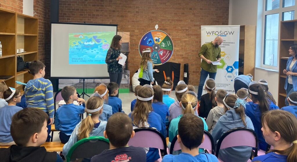 W sali w Sp w Drawsku dzieci siedzą na krzesełkach i słuchają prelekcji prowadzonej przez doradców klimatyczno-energetycznych z WFOŚiGW w Poznaniu.