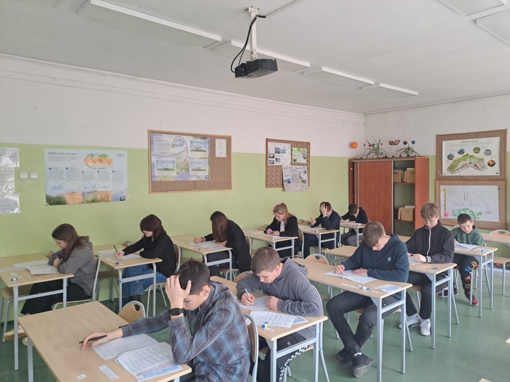 Uczniowie siedzą pojedynczo w ławkach i piszą test w sali.