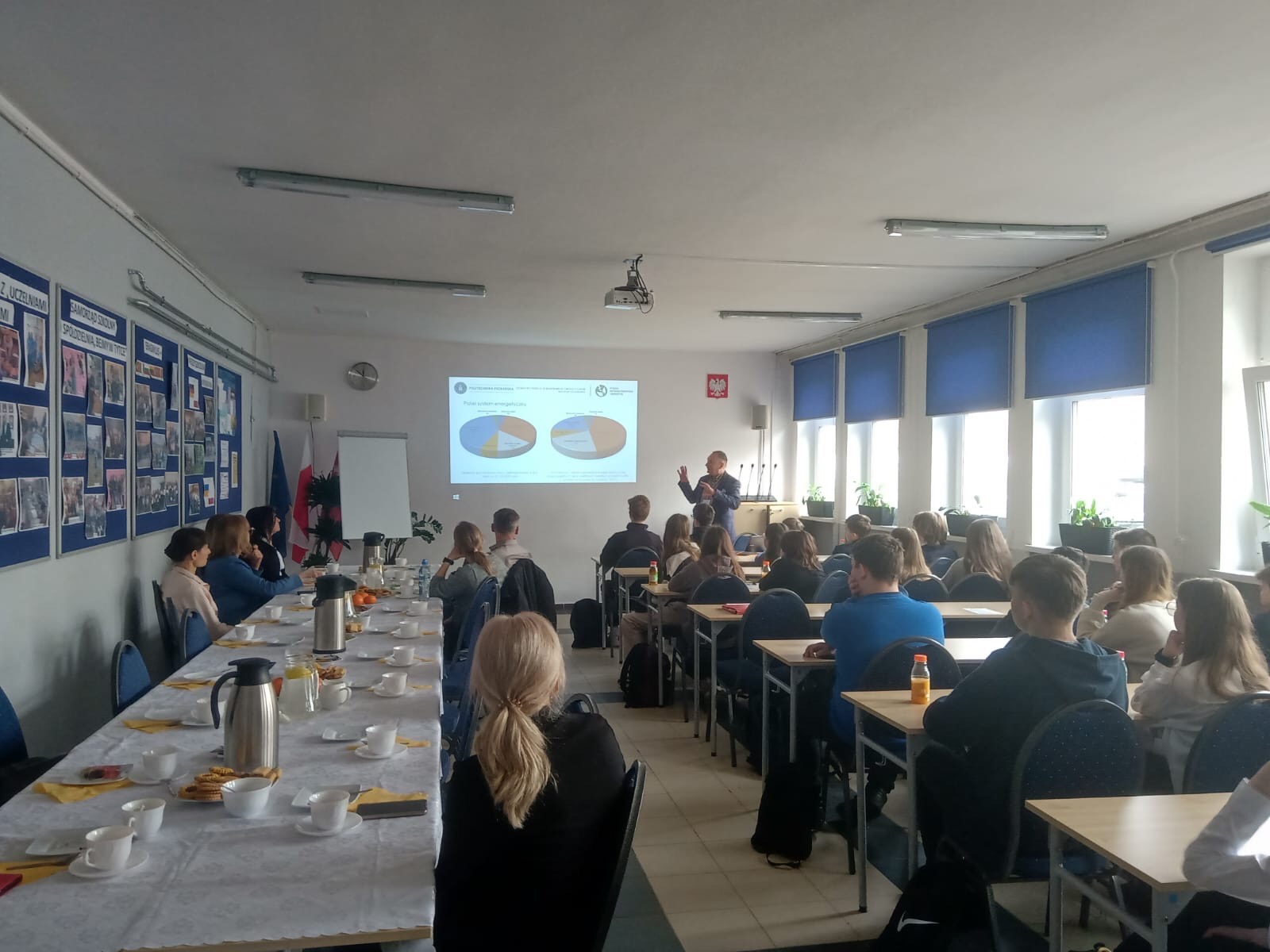 Uczniowie i nauczyciele siedzą w ławkach i słuchają wykładu. Na środku sali stoi jedna osoba i prezentuje przy wyświetlonej na rzutniku prezentacji.