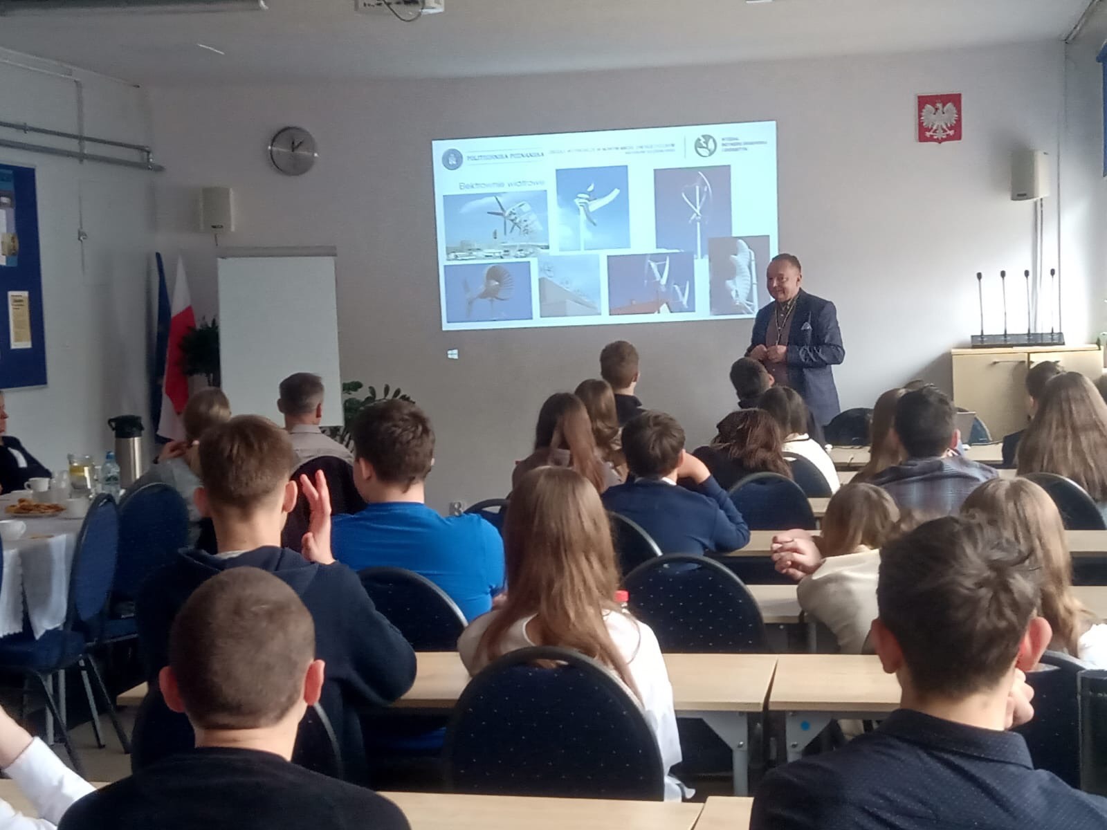 Uczniowie i nauczyciele siedzą w ławkach i słuchają wykładu. Na środku sali stoi jedna osoba i prezentuje przy wyświetlonej na rzutniku prezentacji.