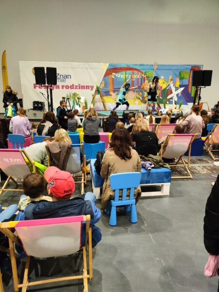 Widok na atrakcje dla dzieci w pobliżu stoiska WFOŚiGW w Poznaniu - gry, pachołki, krzesełka oraz widok na scenę, przed którą siedzi na krzesełkach widownia.