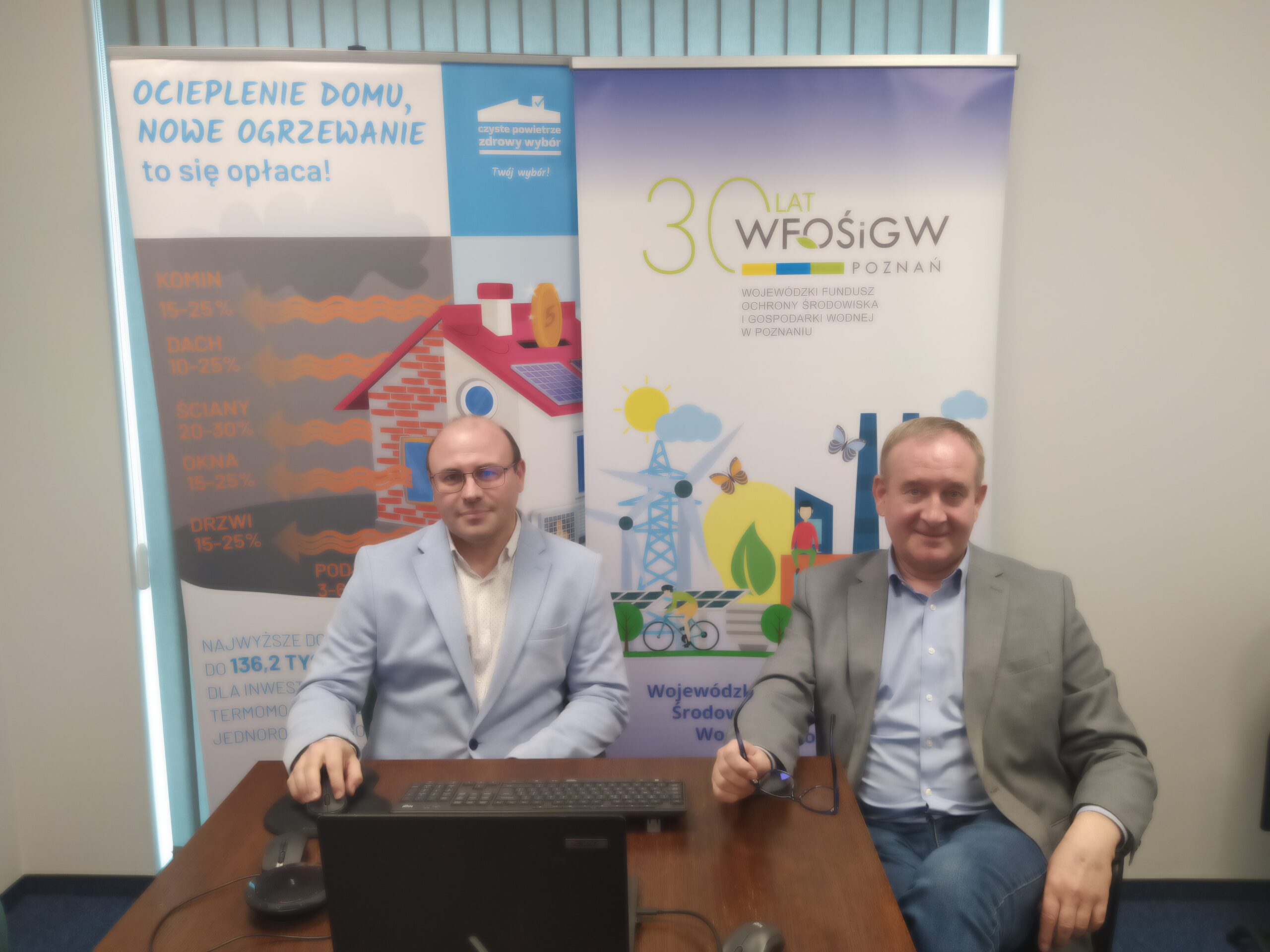 2 osoby siedzą przy stole, na tle bannera reklamowego.