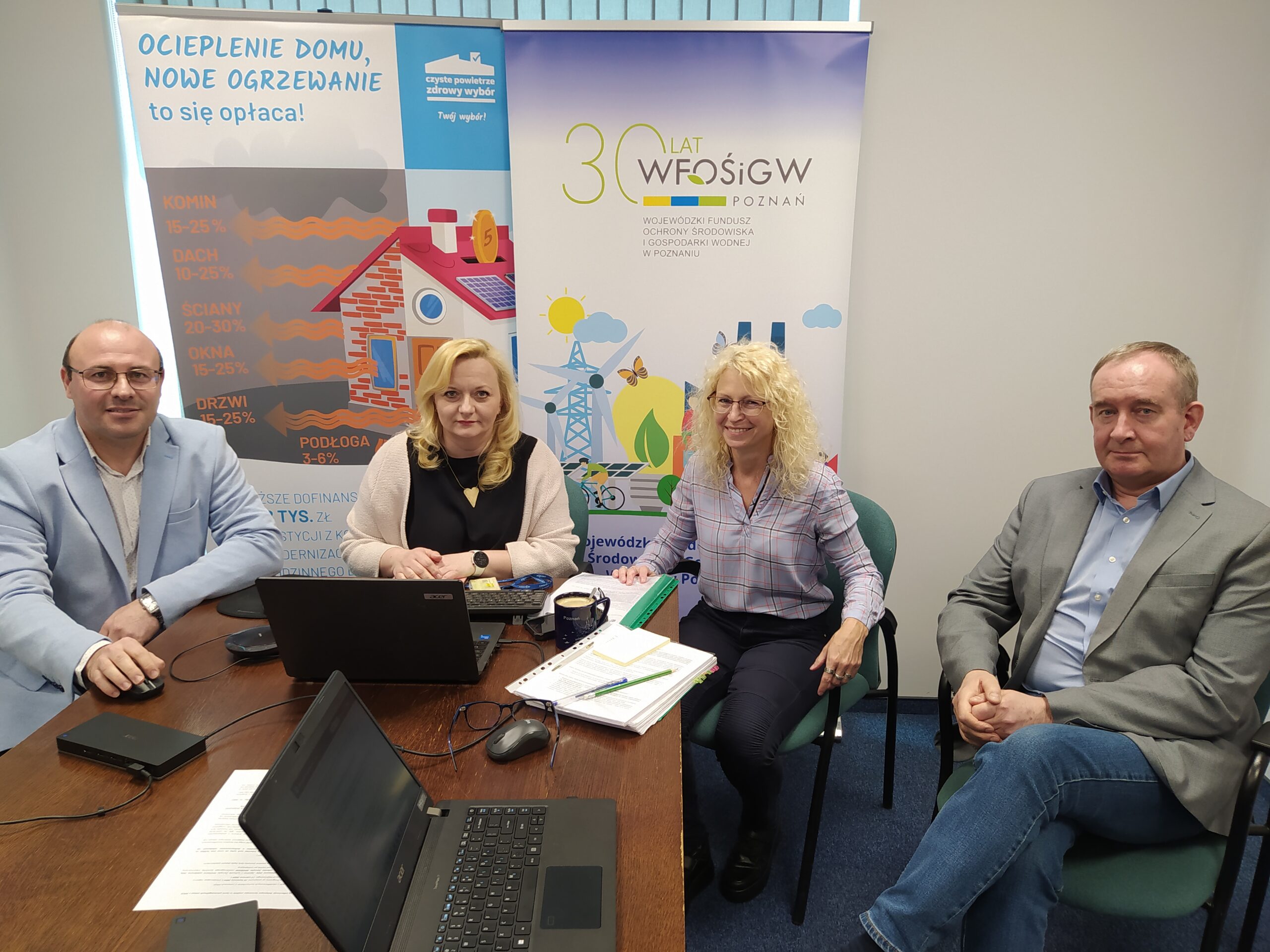 4 osoby siedzą przy stole, na tle bannera reklamowego.