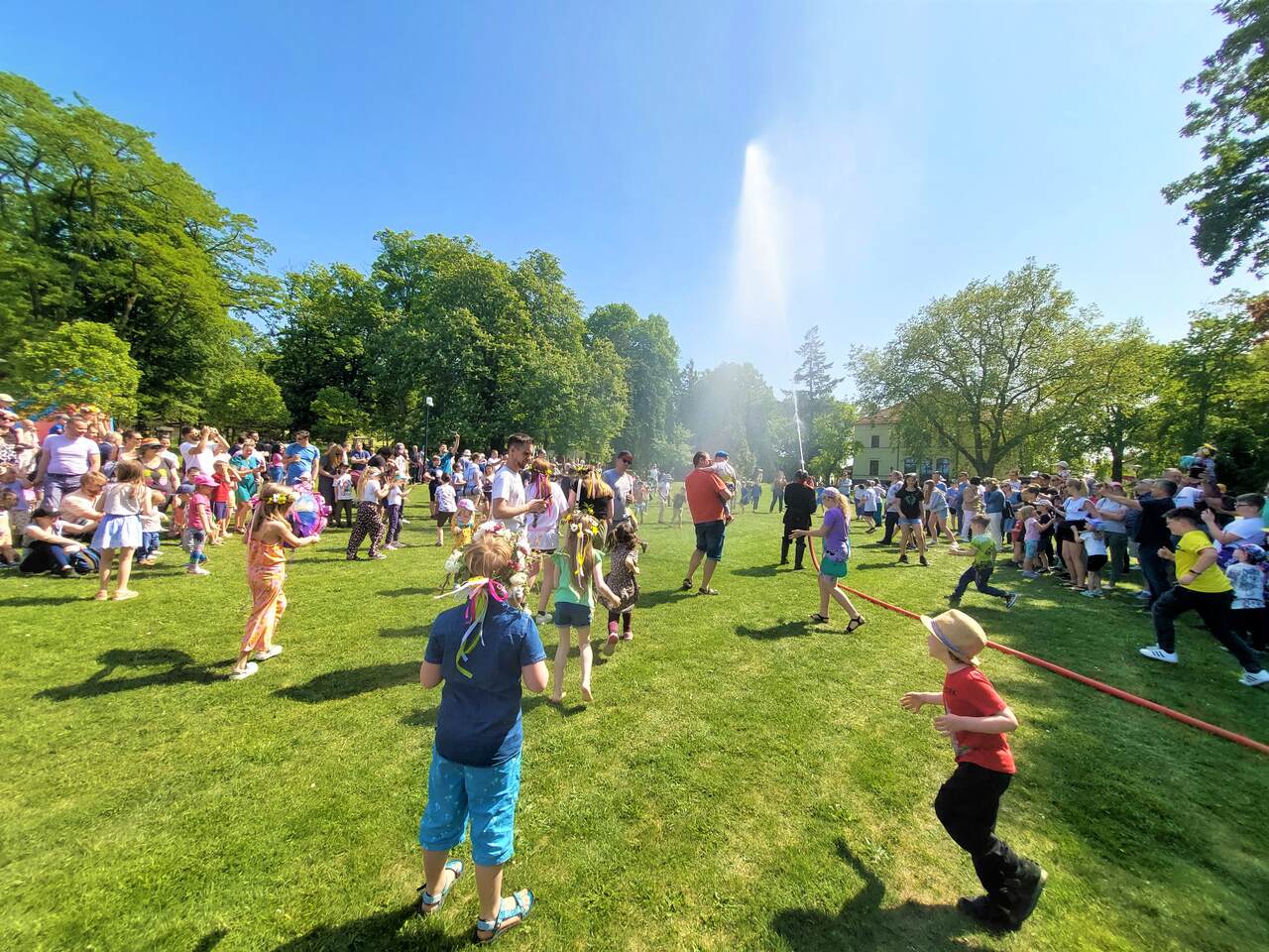 Na terenie festynu z zeszłego roku ludzie spędzają czas na trawiastej powierzchni, bawią się, dzieci biegają pod kurtyną wodną.