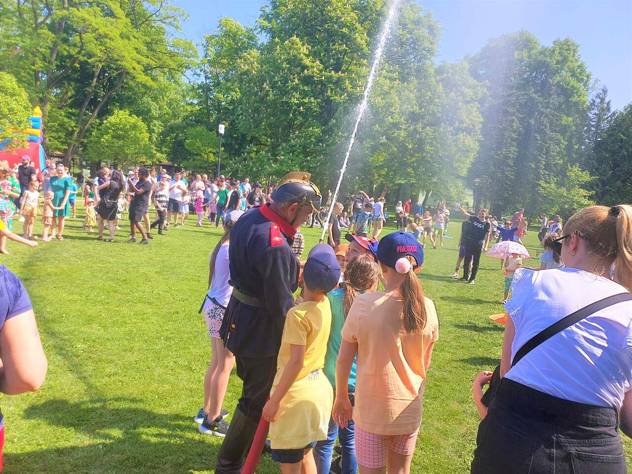 Na terenie festynu z zeszłego roku ludzie spędzają czas na trawiastej powierzchni, bawią się, dzieci biegają pod kurtyną wodną. Strażak w odświętnym tradycyjnym mundurze trzyma wąż z wodą i pomaga dzieciom.