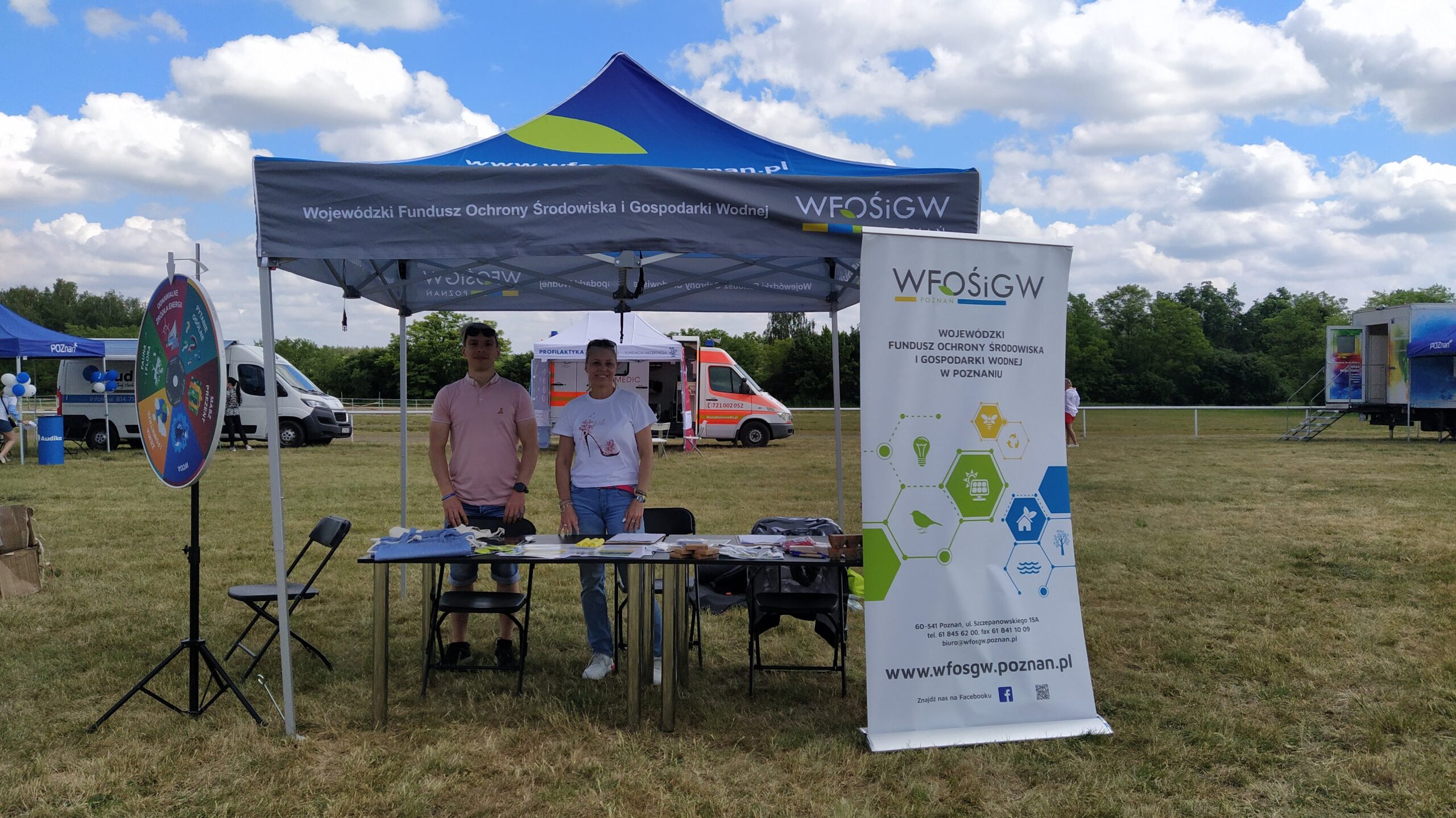 Zdjęcie przedstawia stoisko WFOŚiGW w Poznaniu podczas wydarzenia - namiot wystawienniczy, stół, a za nim 2 przedstawicieli Funduszu- z lewej strony stoi mężczyzna, a z prawej kobieta. Na zdjęciu widoczne jest również koło fortuny.