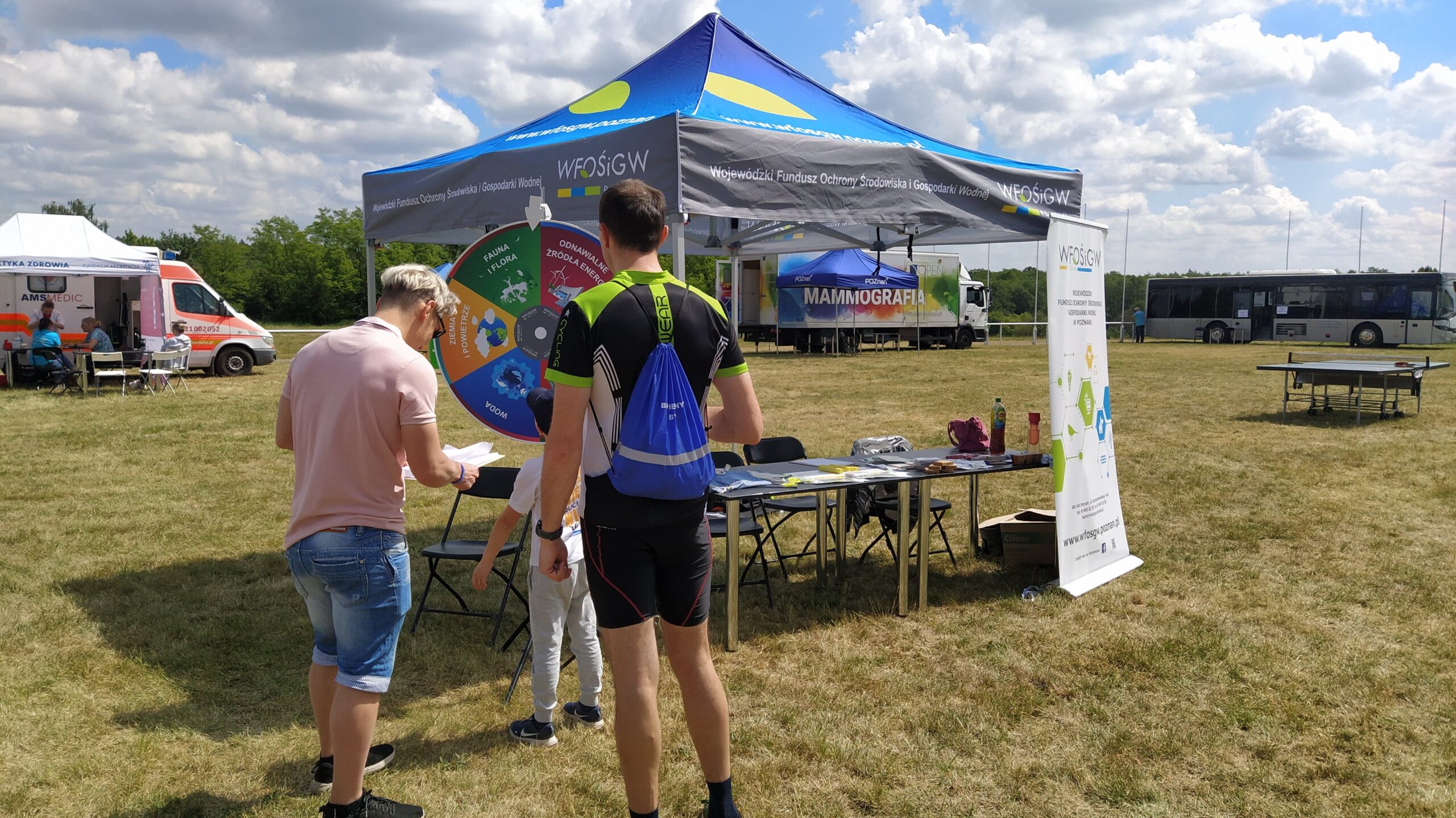Na zdjęciu znajduje się stoisko WFOŚiGW w Poznaniu, a przy stoisku przedstawiciel Funduszu zadający pytanie z koła fortuny małemu chłopcu, obok stoi tata chłopca.