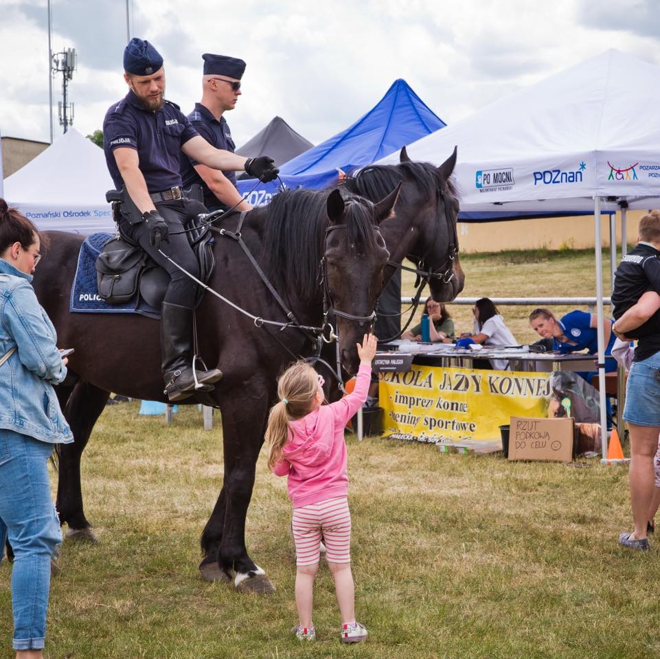 Zdjęcie przedstawia policję konną - dwóchpolicjantów siedzi na koniach, a obok stoi mała dziewczynka, która głaszcze konia, obok stoi jej mama.