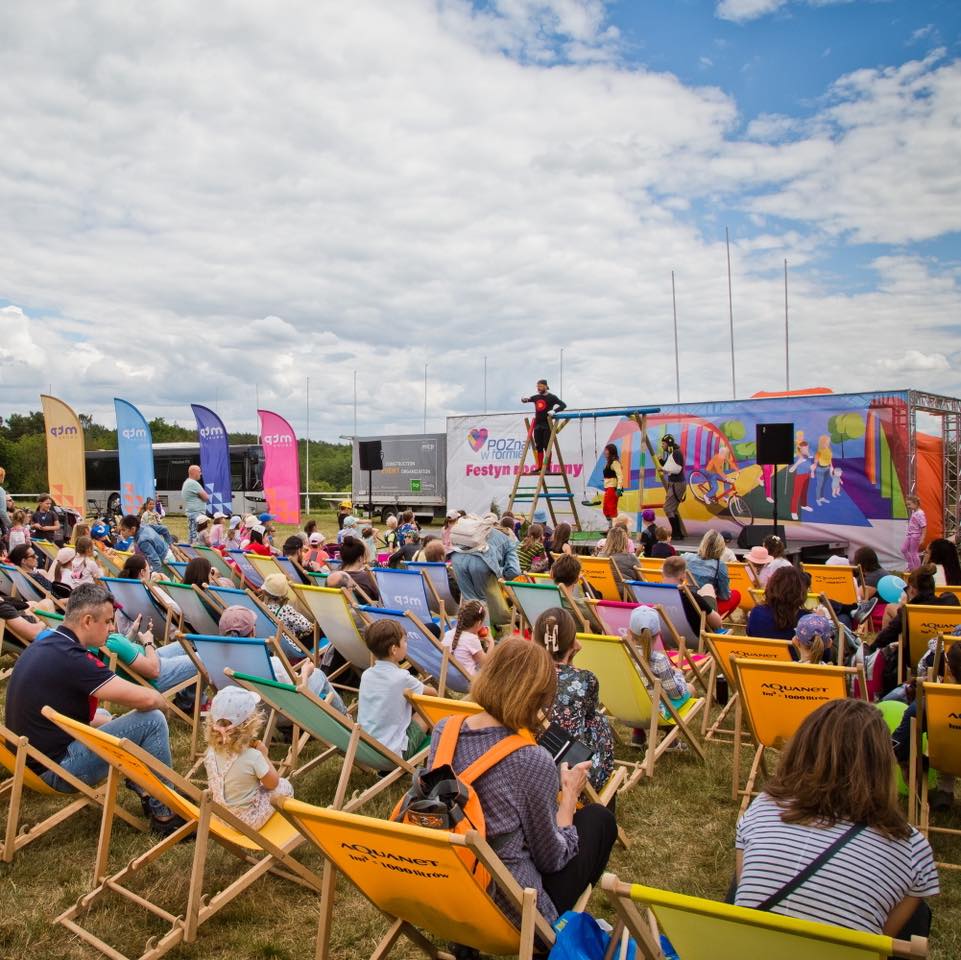Zdjęcie przedstawia grupę ludzi siedzących na leżakach i część stojących wokół, którzy patrzą na występ na scenie podczas festynu.