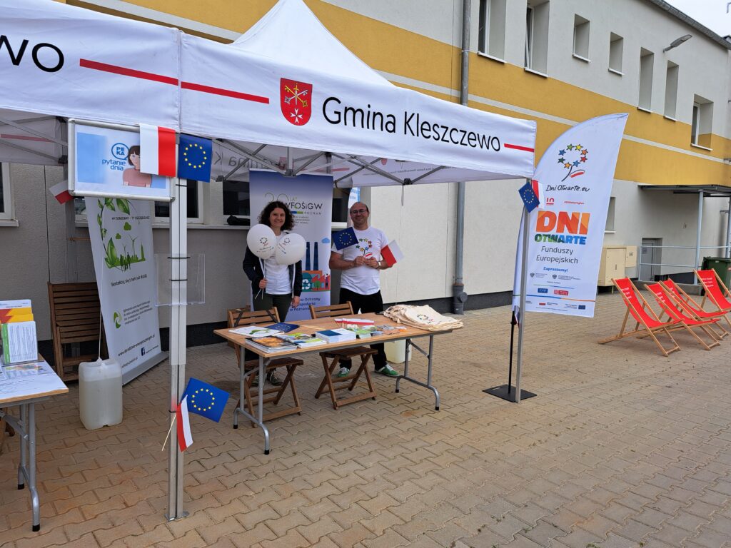 Dwie osoby stoją pod namiotem wystawienniczym za stołem z ulotkami, na tle budynku i bannerów reklamowych WFOŚiGW w Poznaniu.