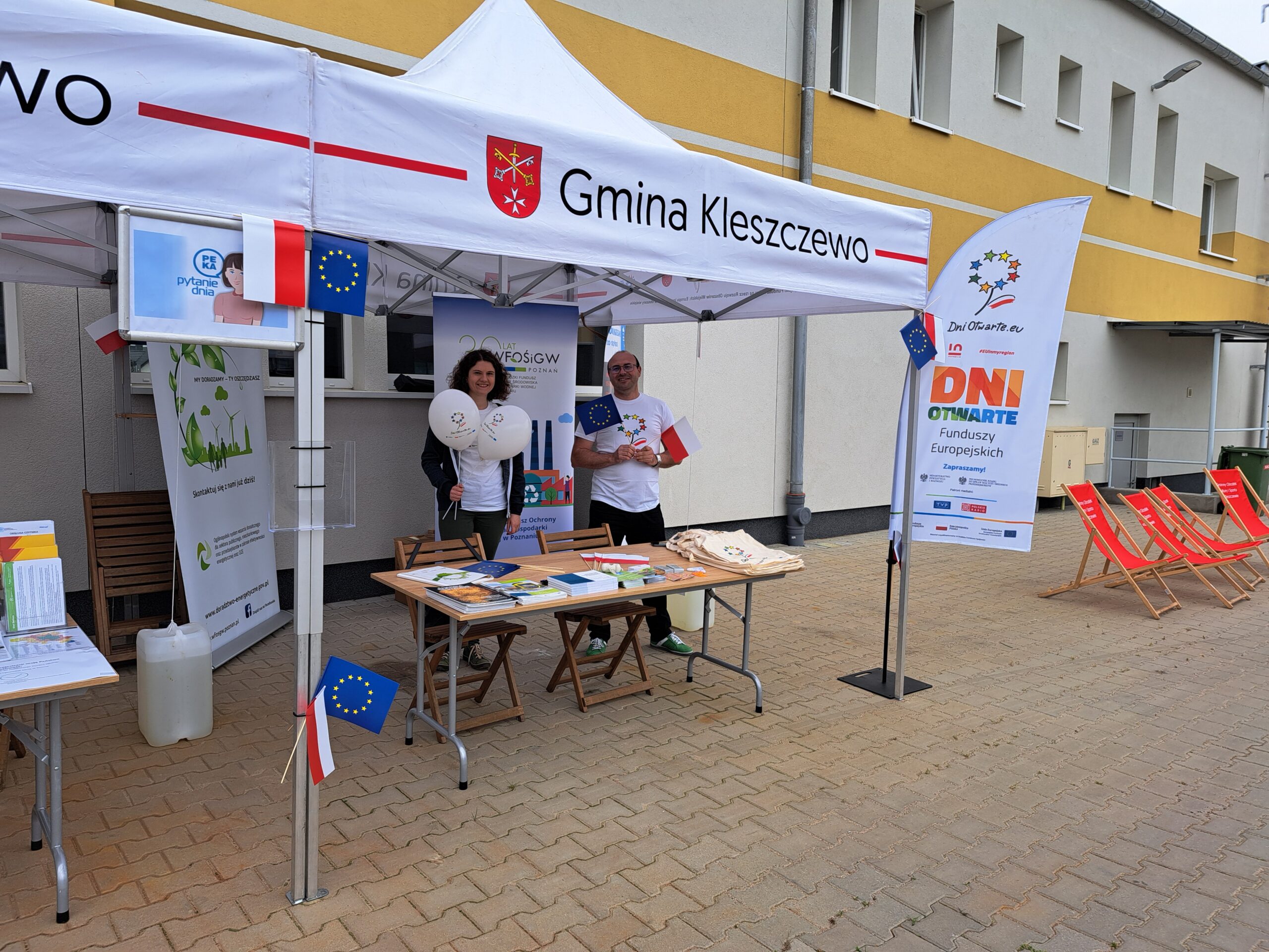 Dwie osoby stoją pod namiotem wystawienniczym za stołem z ulotkami, na tle budynku i bannerów reklamowych WFOŚiGW w Poznaniu.
