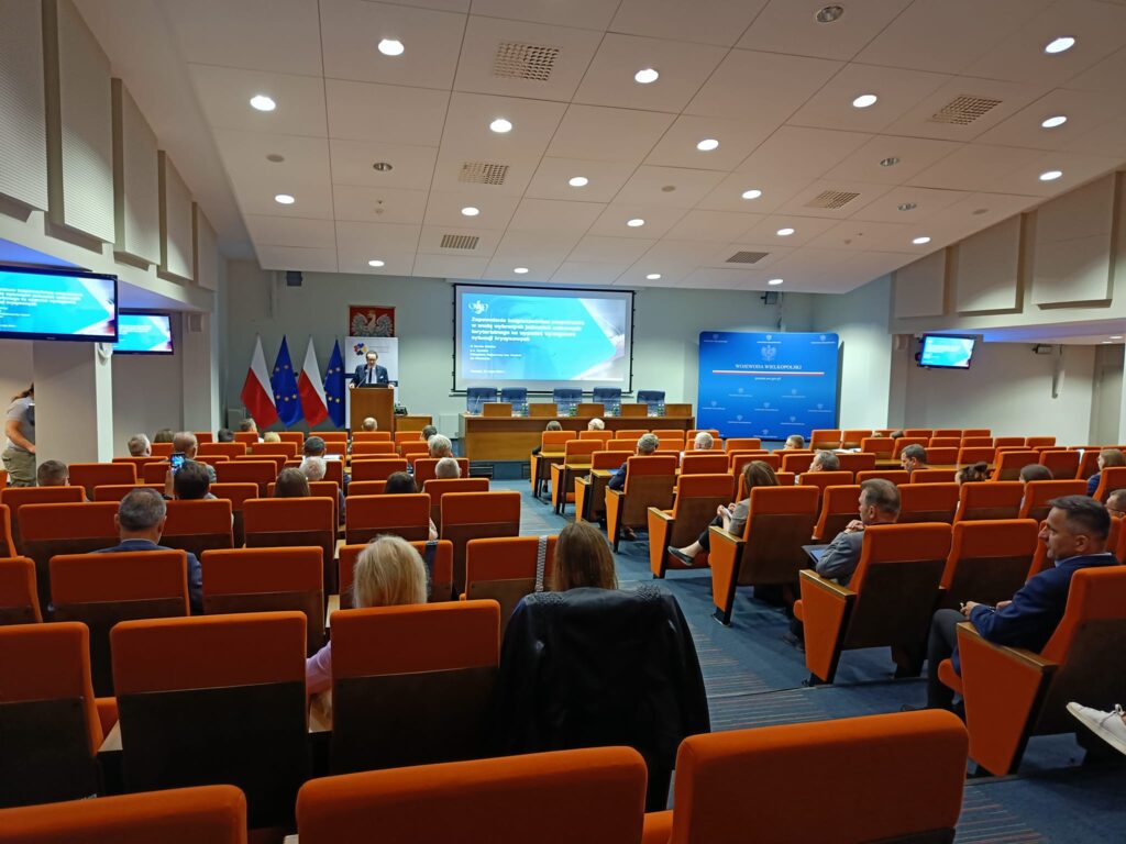 Sala, na której widać osoby siedzące na krzesłach i słuchający prelekcji osoby wygłaszającej jej ze sceny.