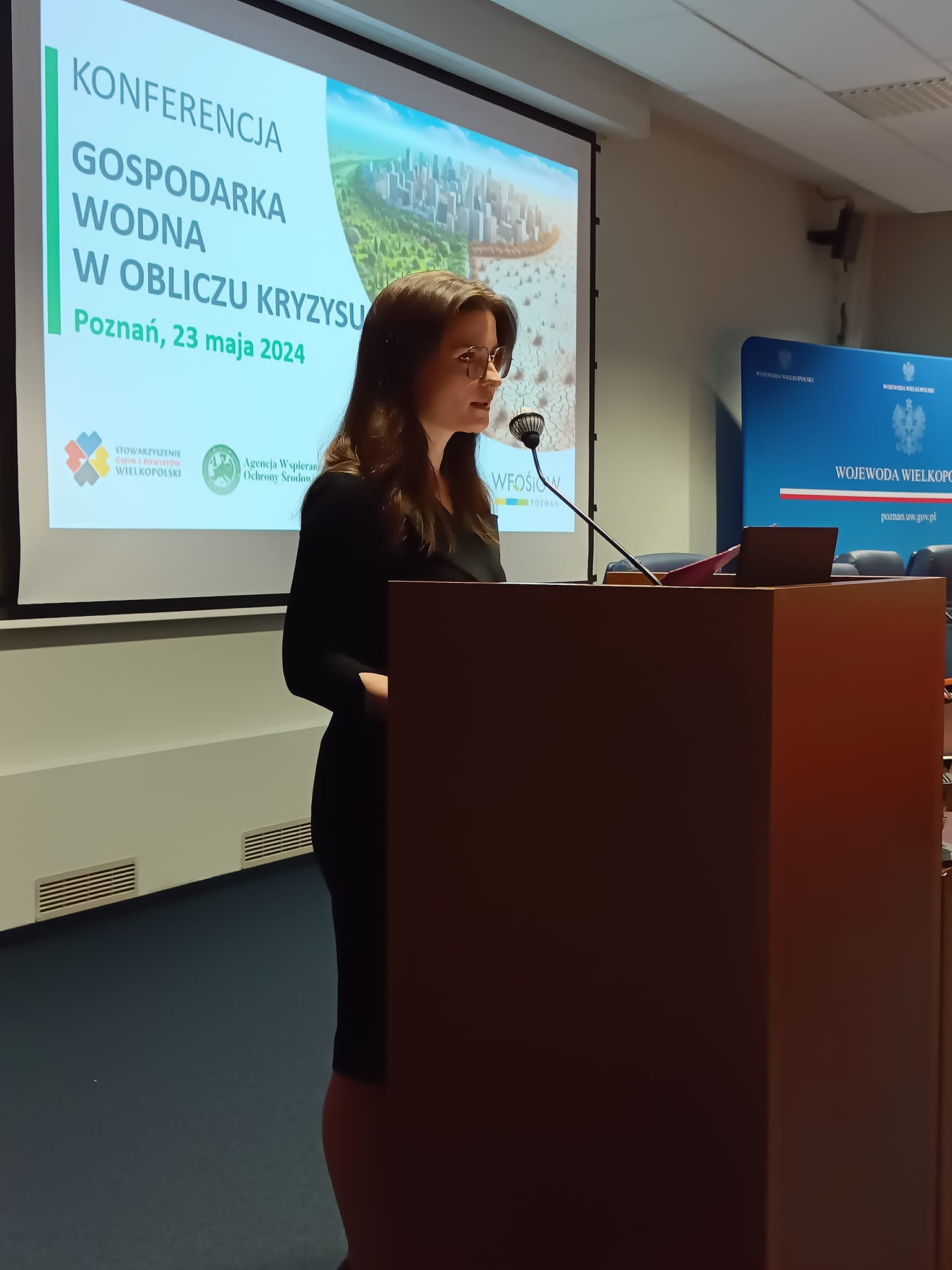 Wojewoda Wielkopolska Agata Sobczak stoi na scenie, na tle wyświetlanej prezentacji i przedstawia prelekcję.