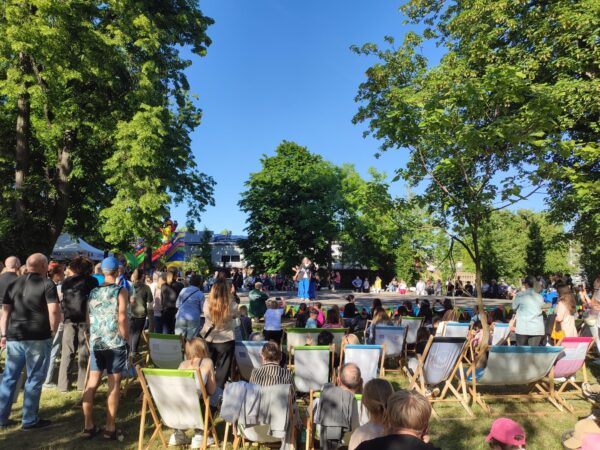 Park, na środku scena, wokół sceny dużo ludzi stojących wokół sceny i siedzących na leżakach.
