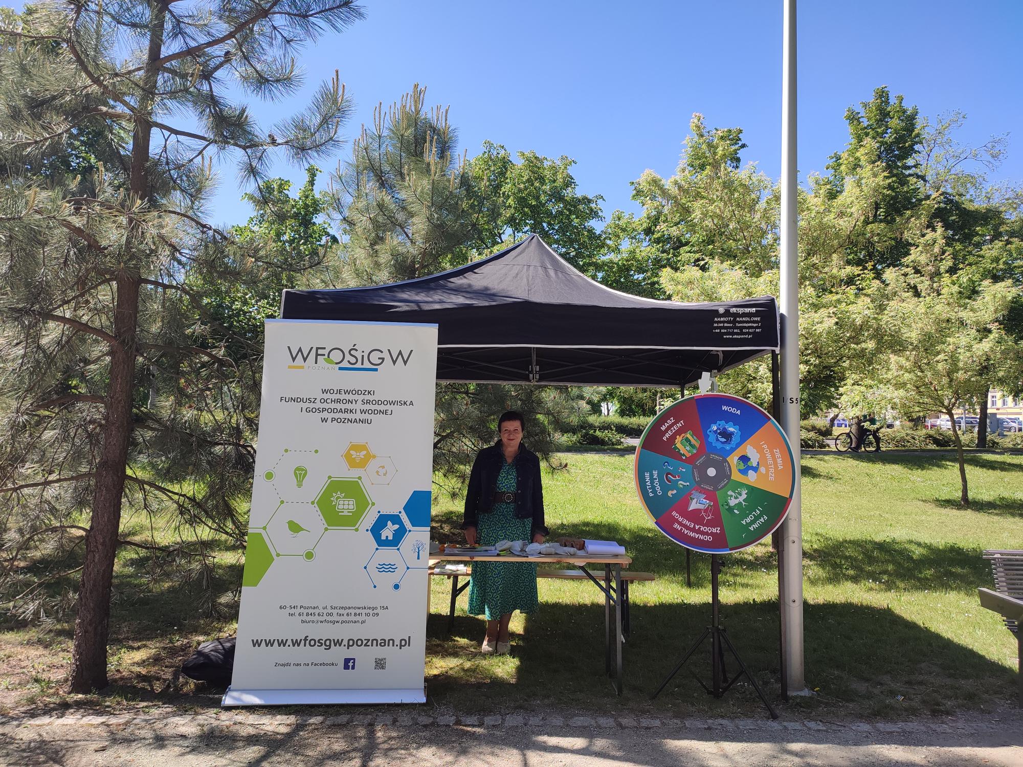 Stoisko WFOŚiGW w parku. Na stoisku stoi jedna osoba za stołem, obok stoiska stoi banner reklamowy i koło fortuny.