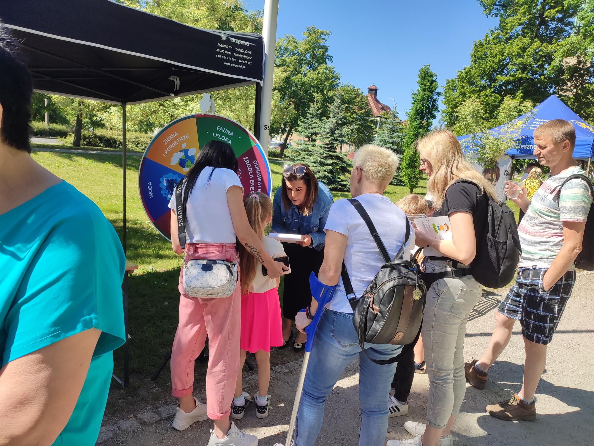 Stoisko WFOŚiGW w parku. Wokół stoiska stoi dużo osób, dzieci biorą udział w zabawie "Ekologiczne koło fortuny".