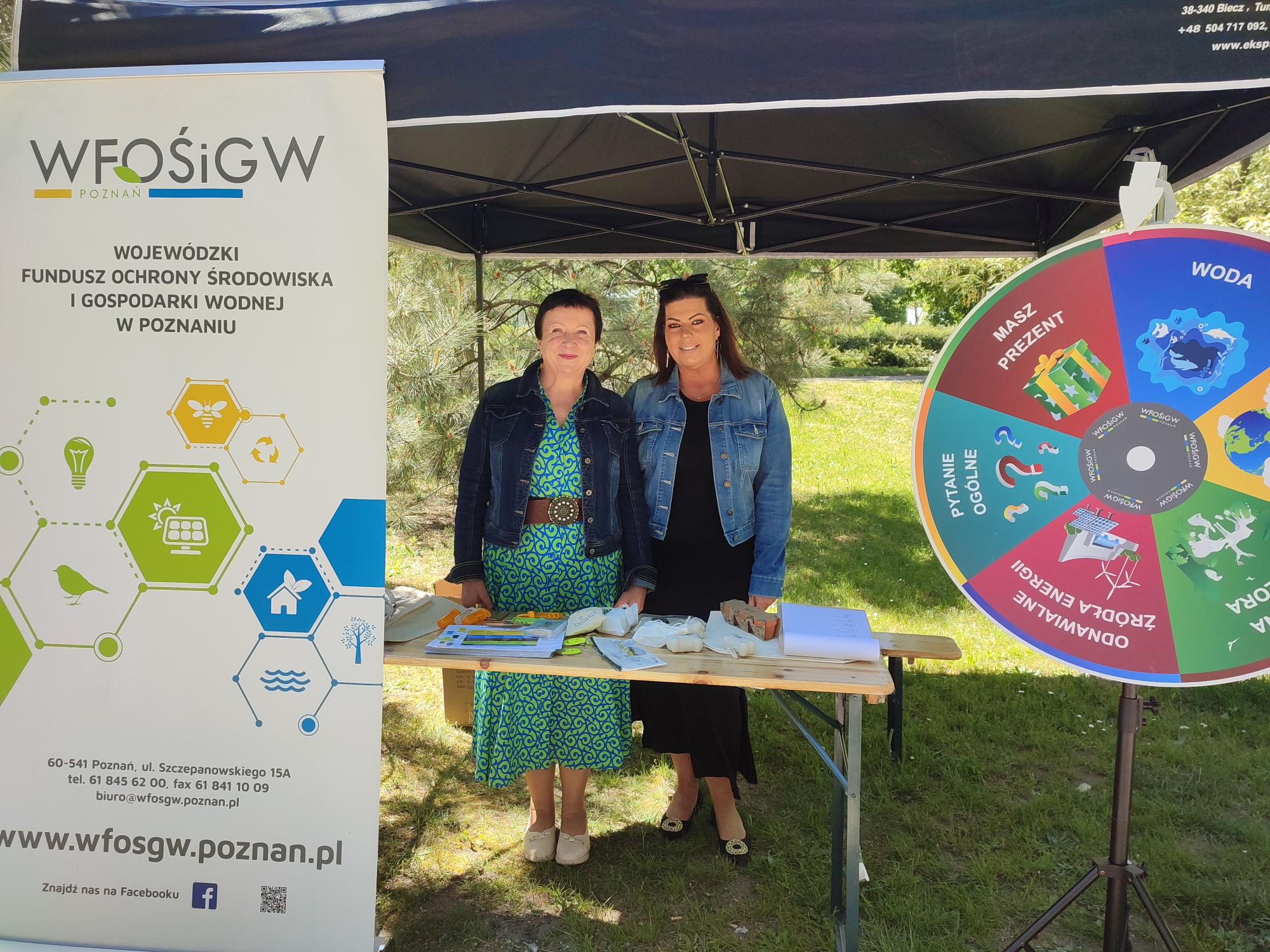 Stoisko WFOŚiGW w Poznaniu, 2 przedstawicielki stoją za stołem,pod namiotem, a przed stoiskiem stoi rollup reklamowy oraz koło fortuny.
