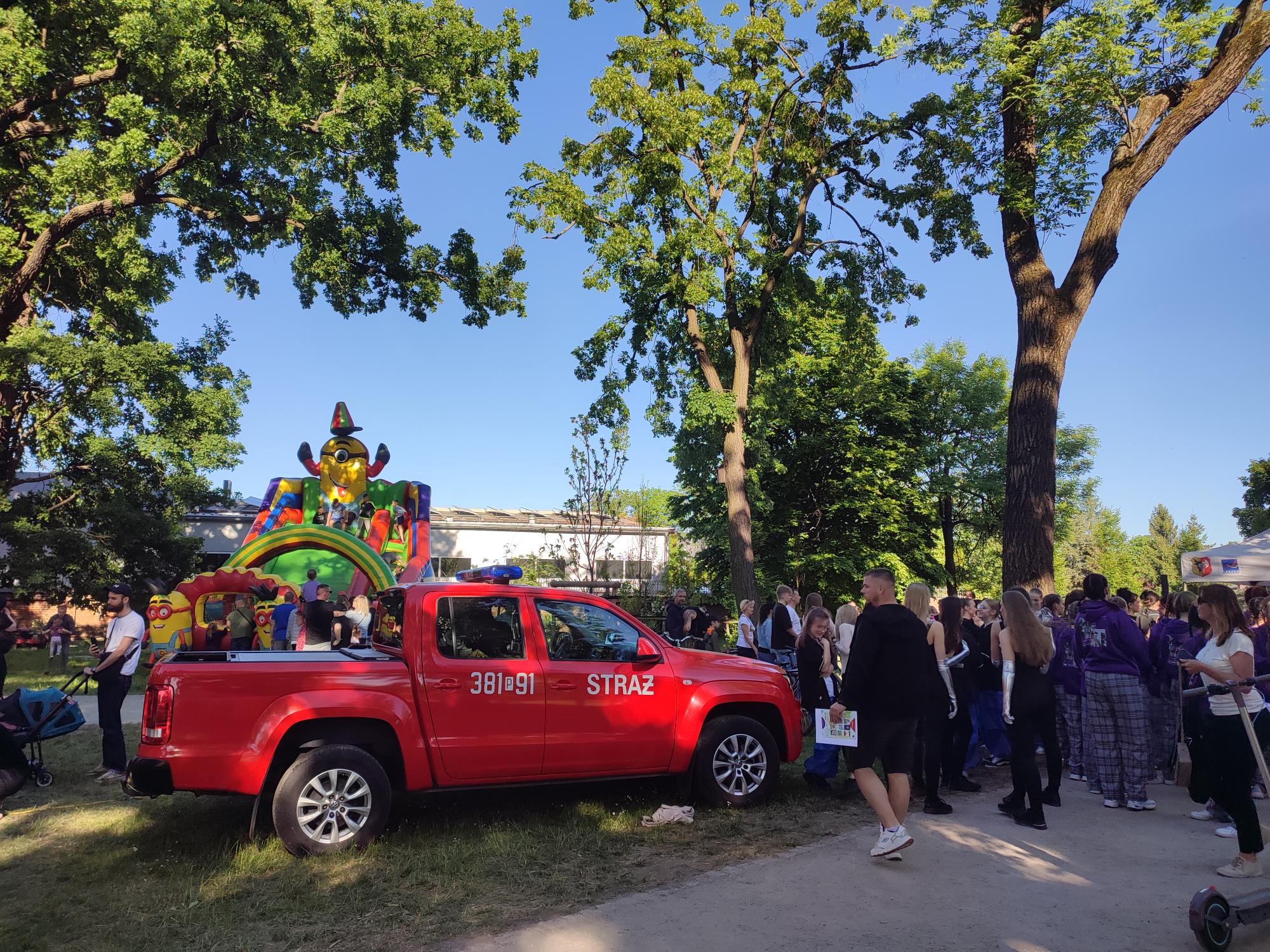 Widok na park, w którym spacerują ludzie podczas pikniku, dzieci bawią się na dmuchańcach. Na pierwszym planie widać jedno z aut straży pożarnej.