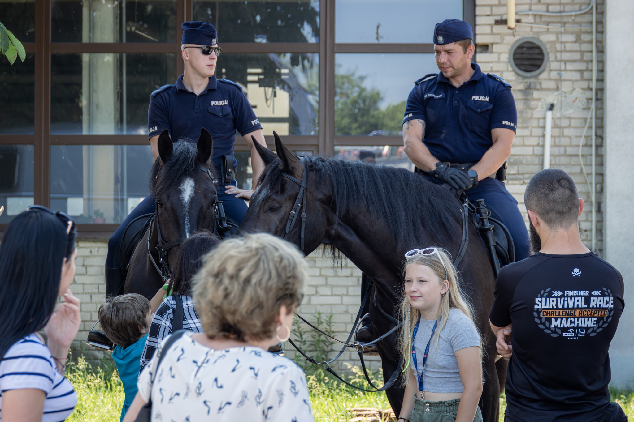 Pokaz policji konnej, na placu dwóch policjantów na koniach, opowiada o koniach zgromadzonym dzieciom, wokół zgromadzeni są dorośli i dzieci.
