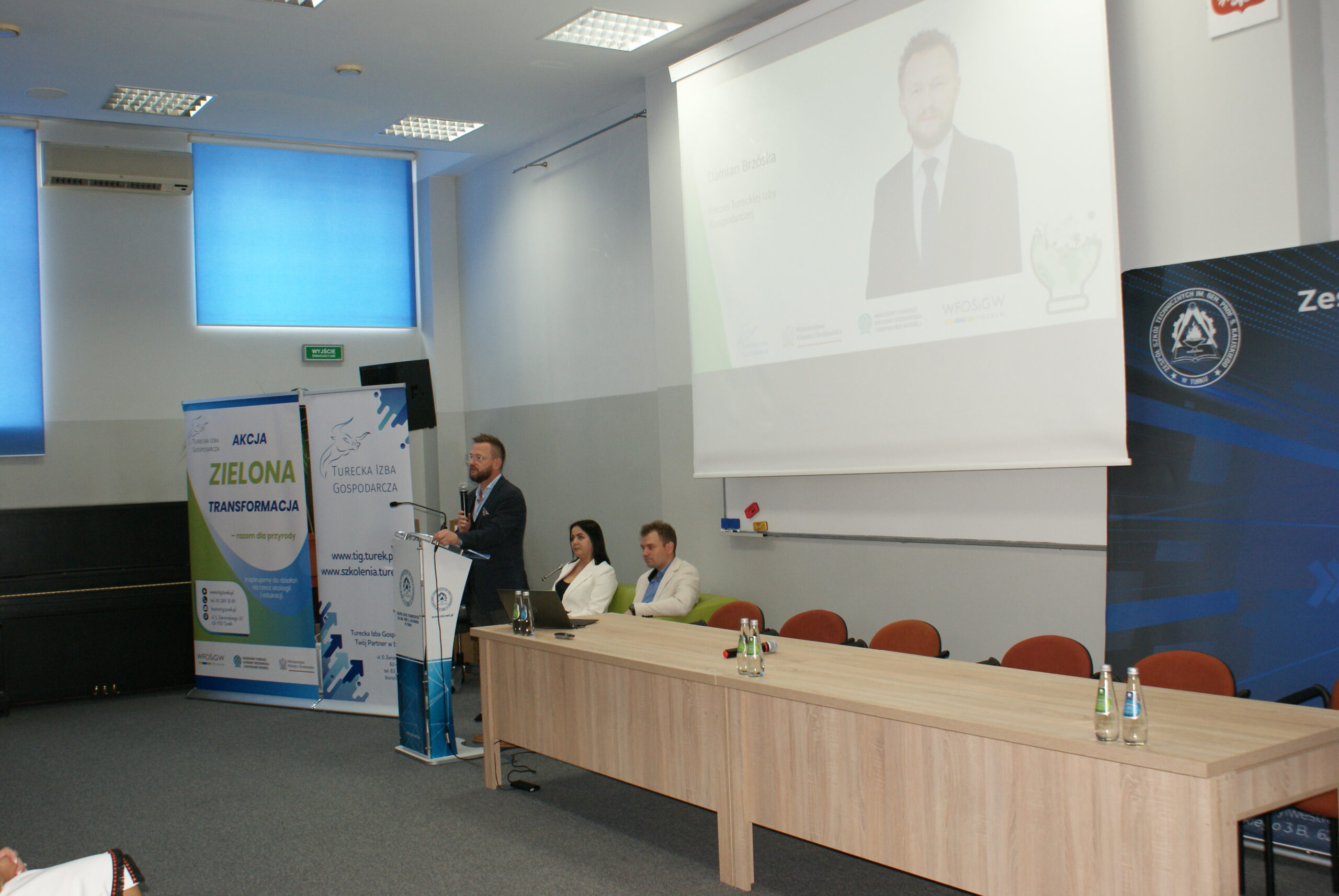 1osoba stoi na scenie i przemawia, w tle prezentacja.