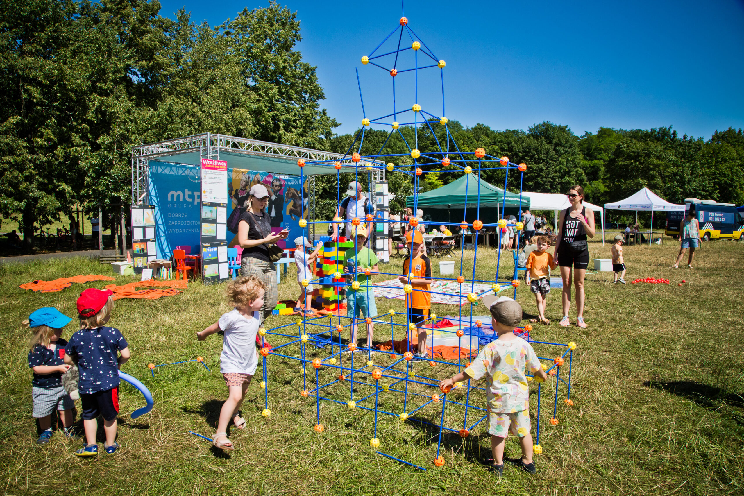 Zdjęcie przedstawia dzieci bawiące sie podczas festynu, tutaj grupa dzieci układa przestrzenne konstrukcje z klocków pod okiem animatorów.