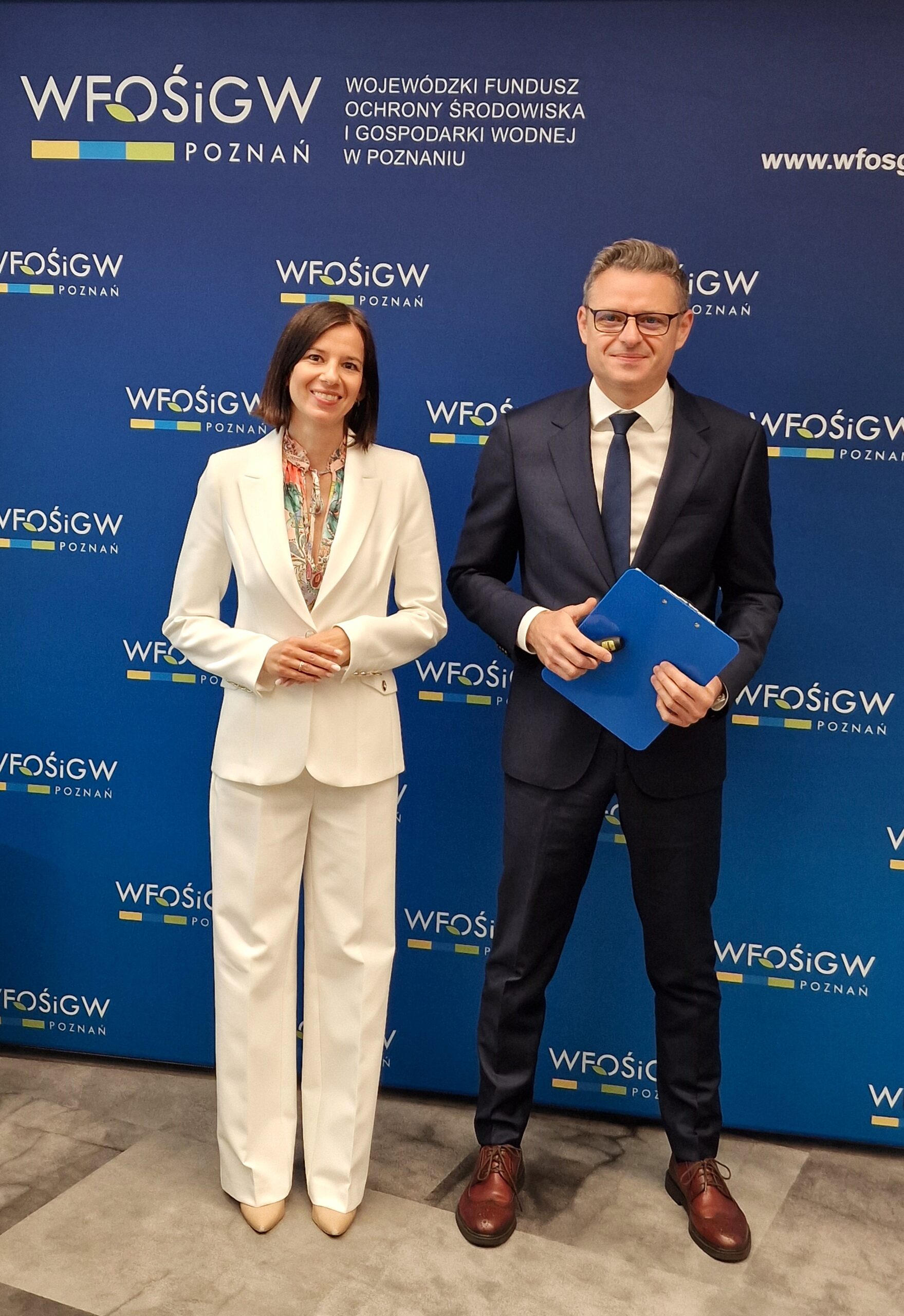 Dwie osoby - Zarząd WFOŚiGW w Poznaniu - stoją na tle ścianki reklamowej z logotypami.
