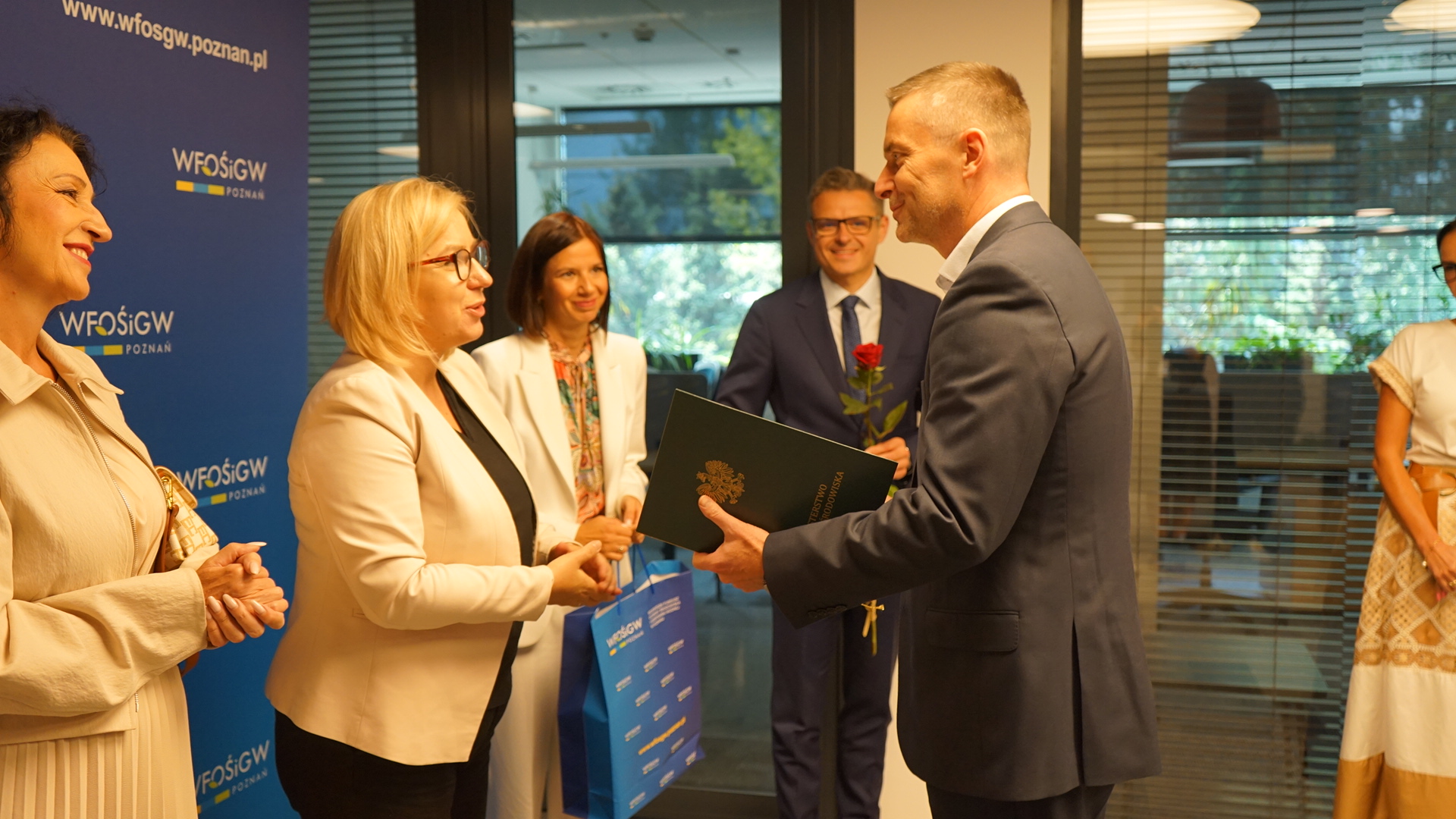 Przy ściance reklamowej stoi kilka osób, jednej osobie wręczany jest list gratulacyjny, torba z upominkami i kwiat.