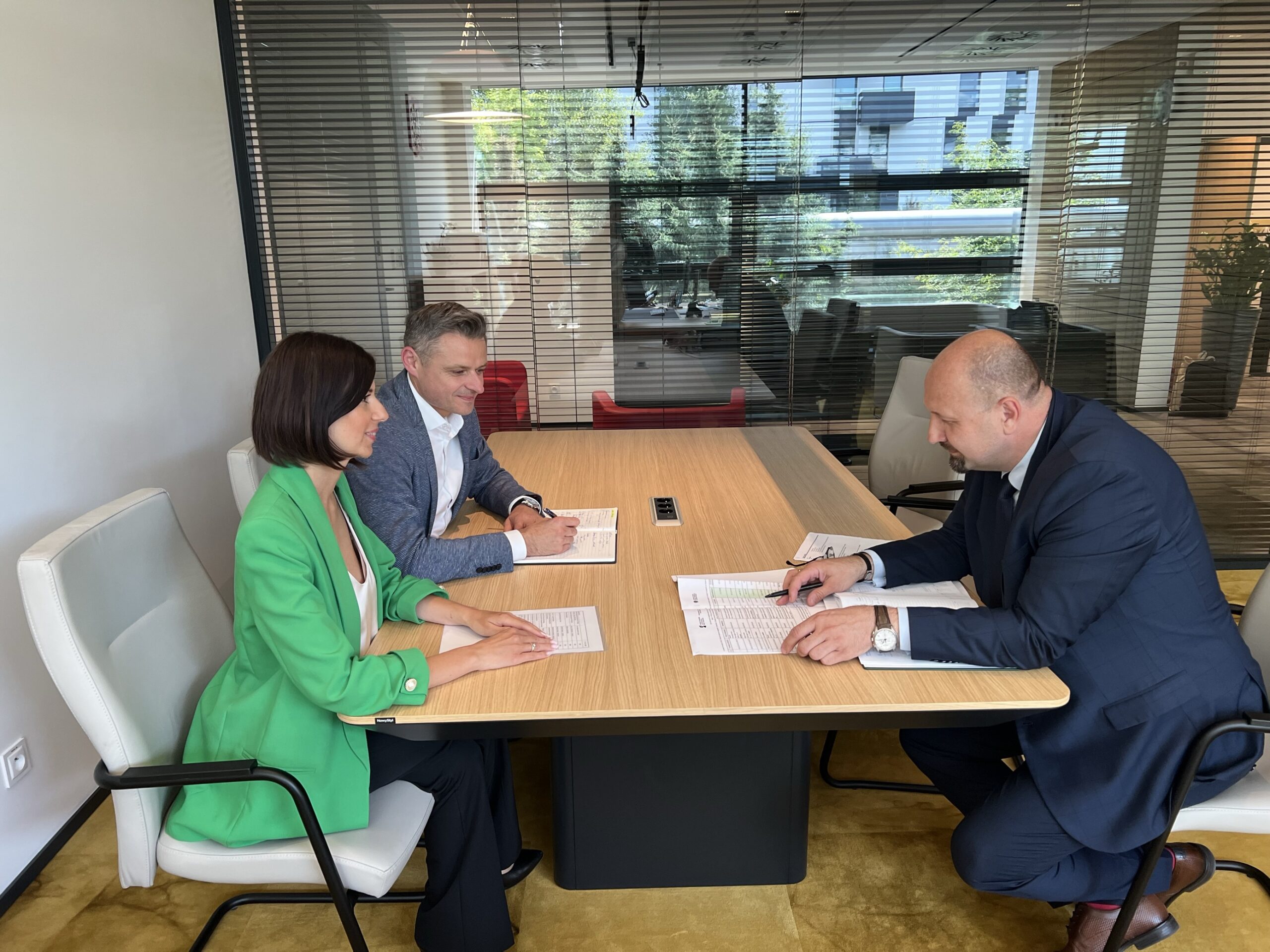 W biurze, w gabinecie Prezesa, przy stole siedzą 3 osoby, po lewej stronie Prezes Zarządu WFOŚiGW w Poznaniu wraz z Zastępcą Prezesa Zarządu oraz naprzeciwko, po prawej stronie zdjęcia Zastępca Prezesa Zarządu NFOŚiGW. Przed nimi na stole leżą dokumenty, a osoby rozmawiają.