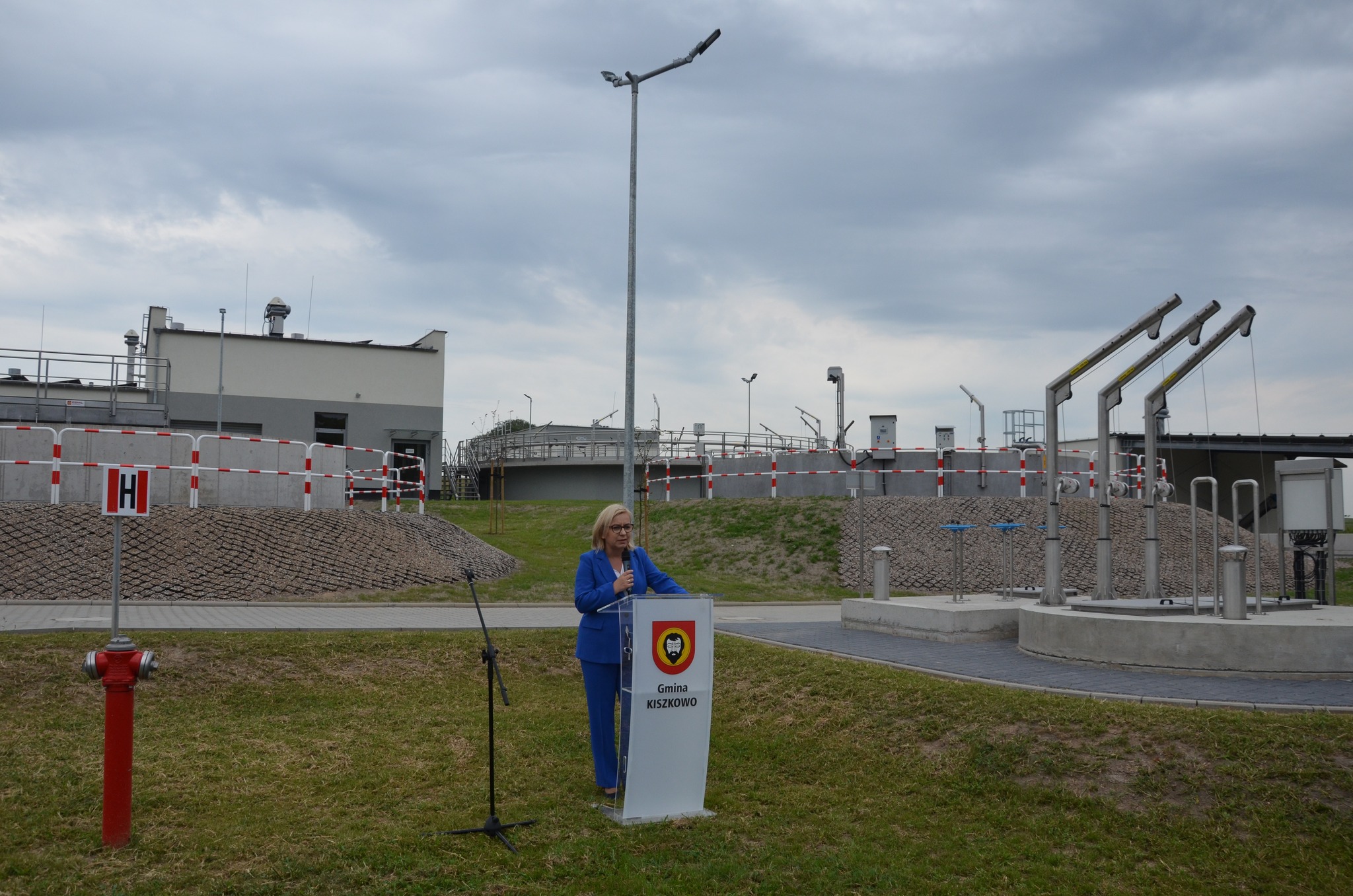 Na zdjęciu na tle budynku oczyszczalni ścieków stoi przy mównicy Minister Klimatu i Środowiska i przemawia.