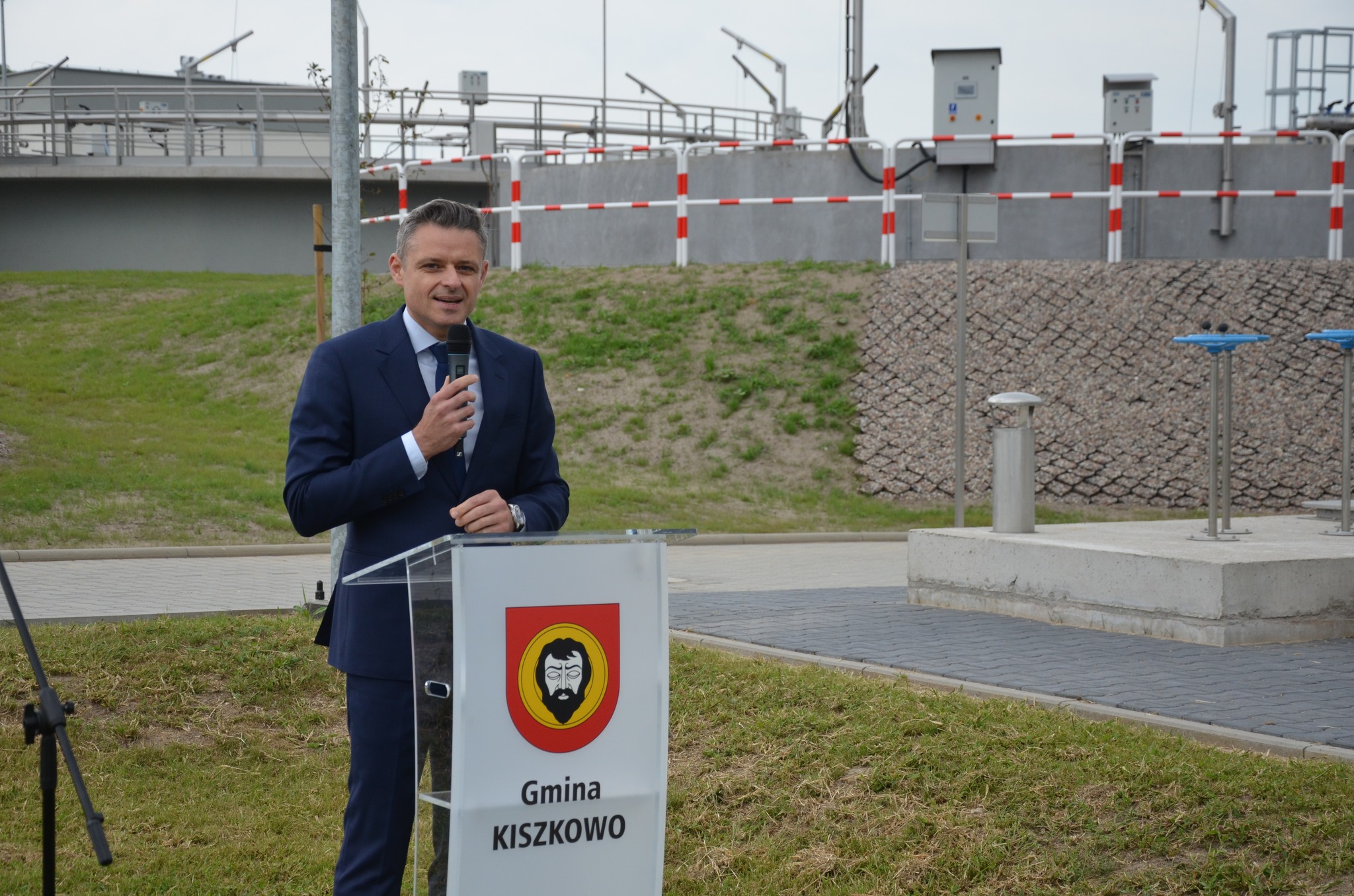 Na zdjęciu na tle budynku oczyszczalni ścieków stoi przy mównicy Prezes Zarządu WFOŚiGW w Poznaniu, Pan Rafał Ratajczak, i przemawia.