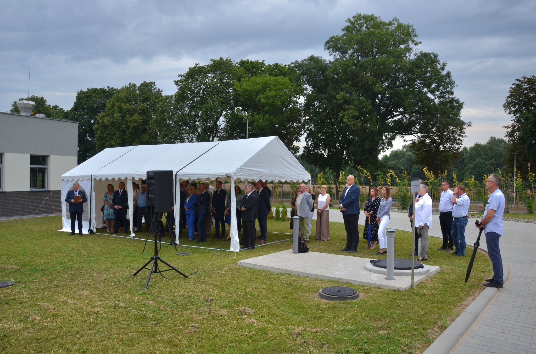 Na zdjęciu obok zmodernizowanej oczyszczalni ścieków, na trawie stoi namiot, a pod nim zgromadzeni goście uroczystości.