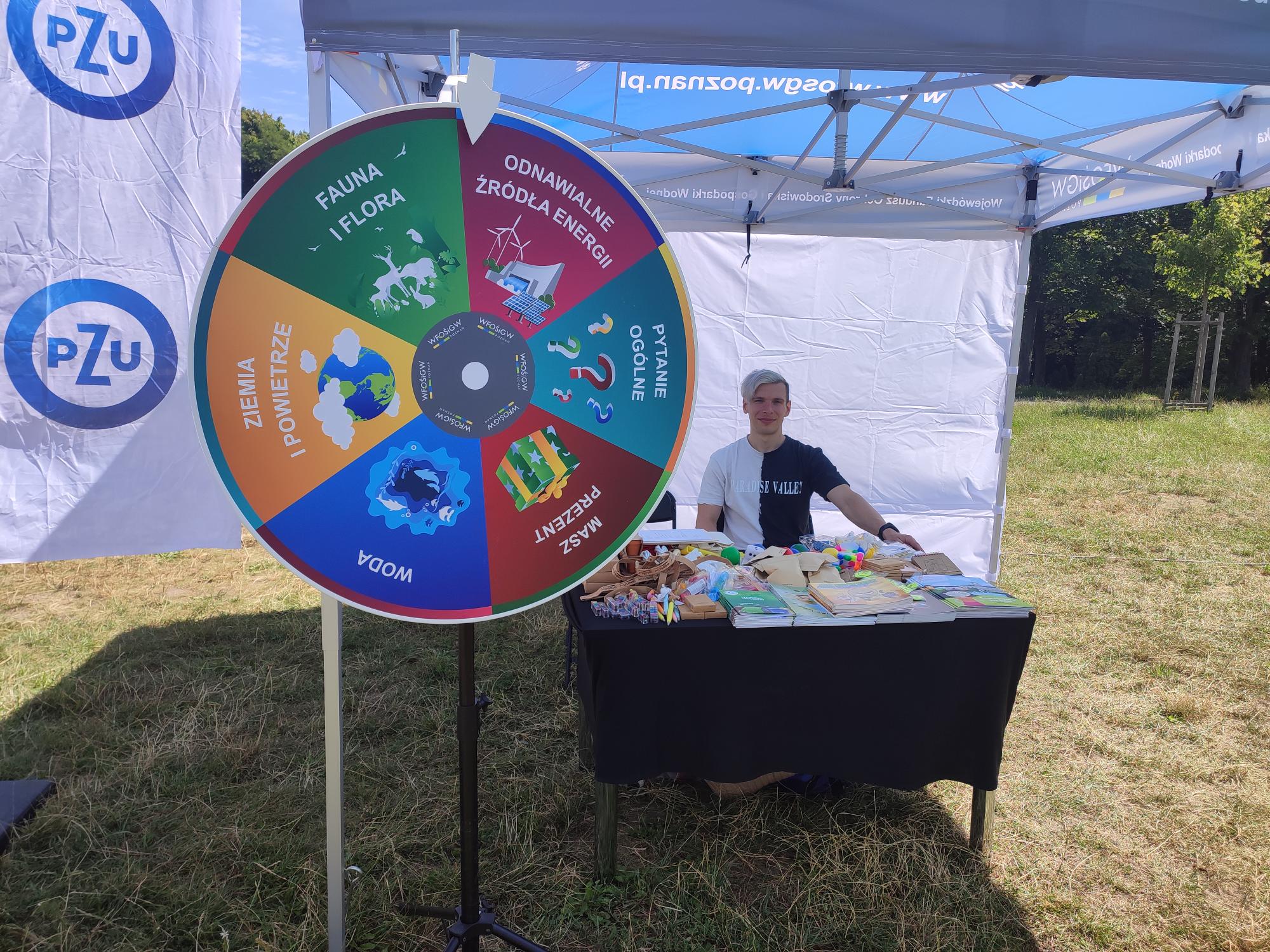 Na zdjęciu widać stoisko WFOŚiGW w Poznaniu - pod namiotem stół oraz jedna z osób obsługujących stoisko, tuż przed namiotem stoi koło fortuny.