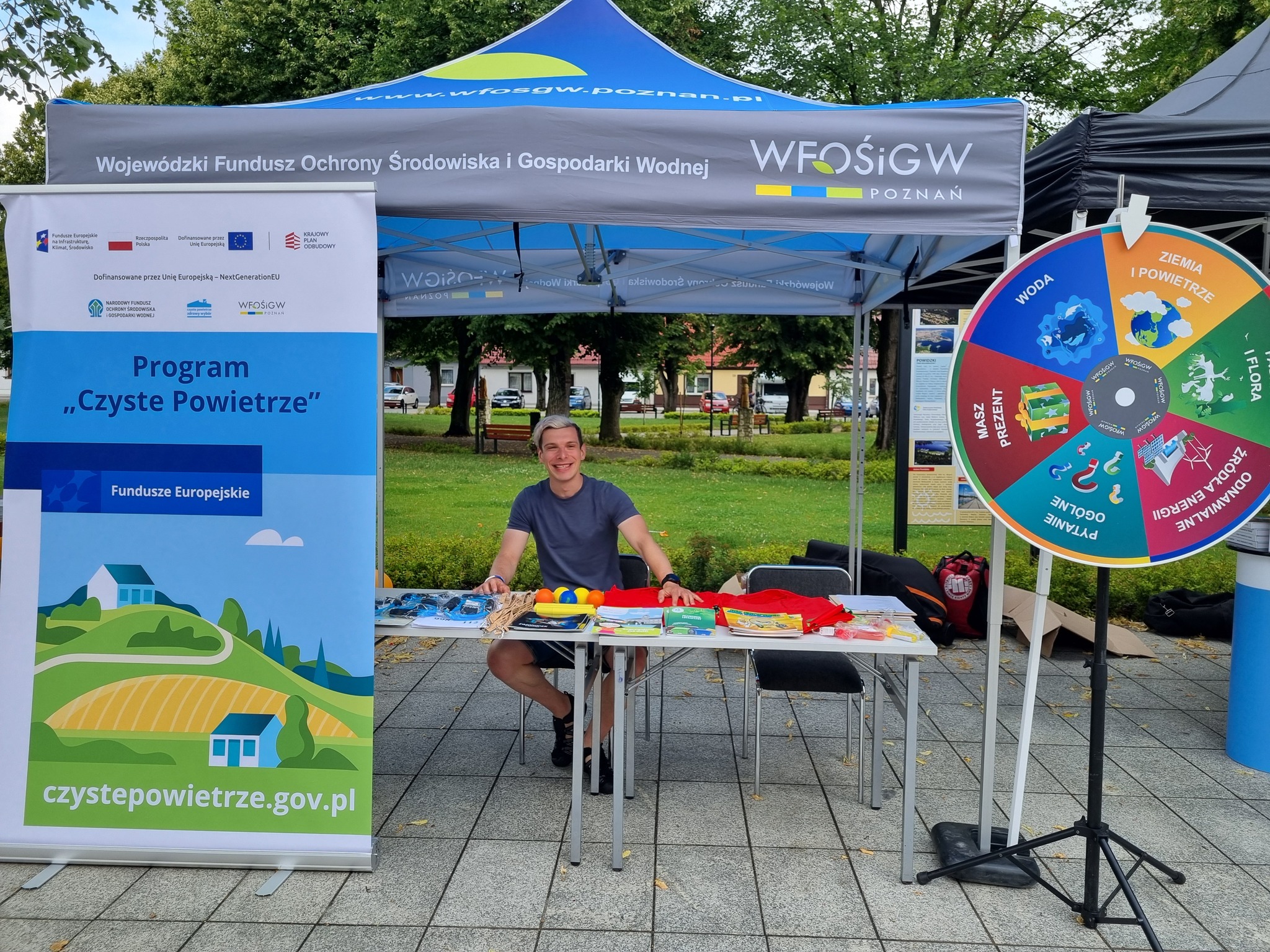 Zdjęcie przedstawia stoisko WFOŚiGW w Poznaniu - namiot, a pod nim przystoliku siedzi pracownik, przed stoiskiem widać rollup reklamowy i koło fortuny.