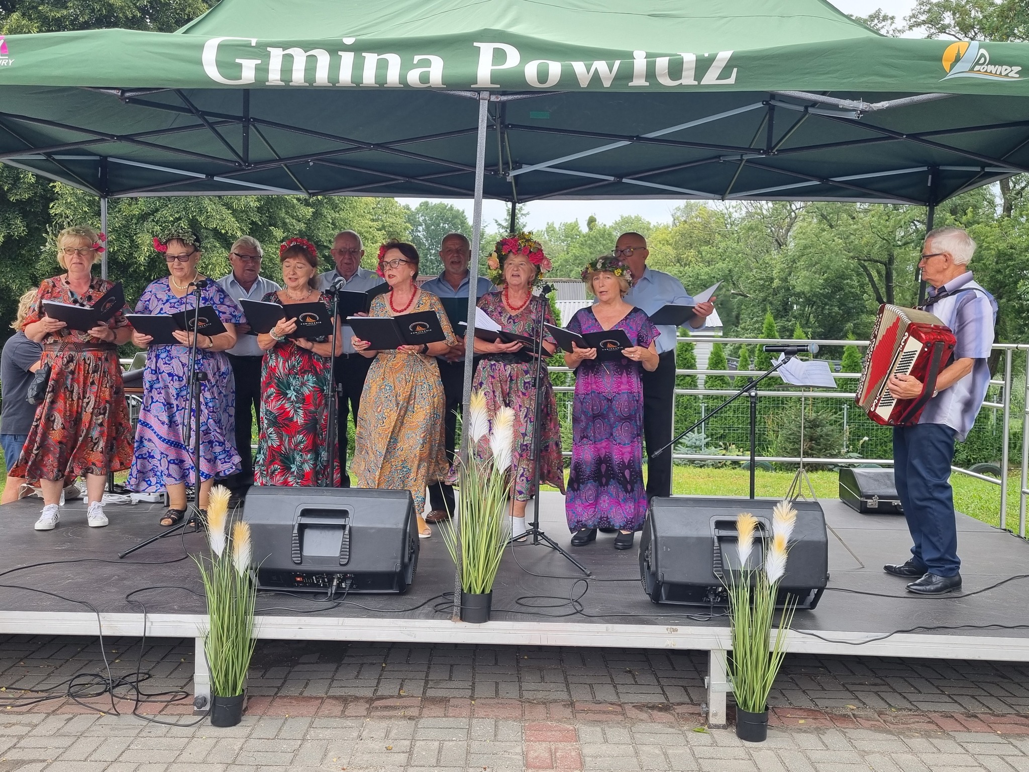 Zdjęcie przedstawia scenę i występ zespołu grupa osób stoi na scenie ze śpiewnikami i śpiewa, obok stoi pan, który akompaniuje na akordeonie.