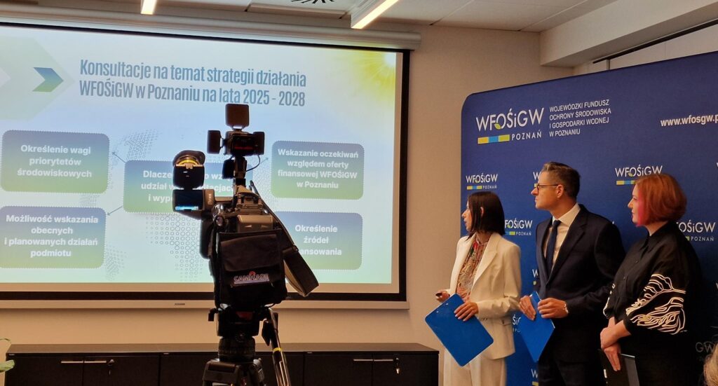 Zdjęcie przedstawia widok na wyświetlana prezentacja, a po prawej stronie, na tle ścianki z logotypami WFOŚiGW stoją 3 osoby i przemawiają. Na pierwszym planie widoczna jest kamera.