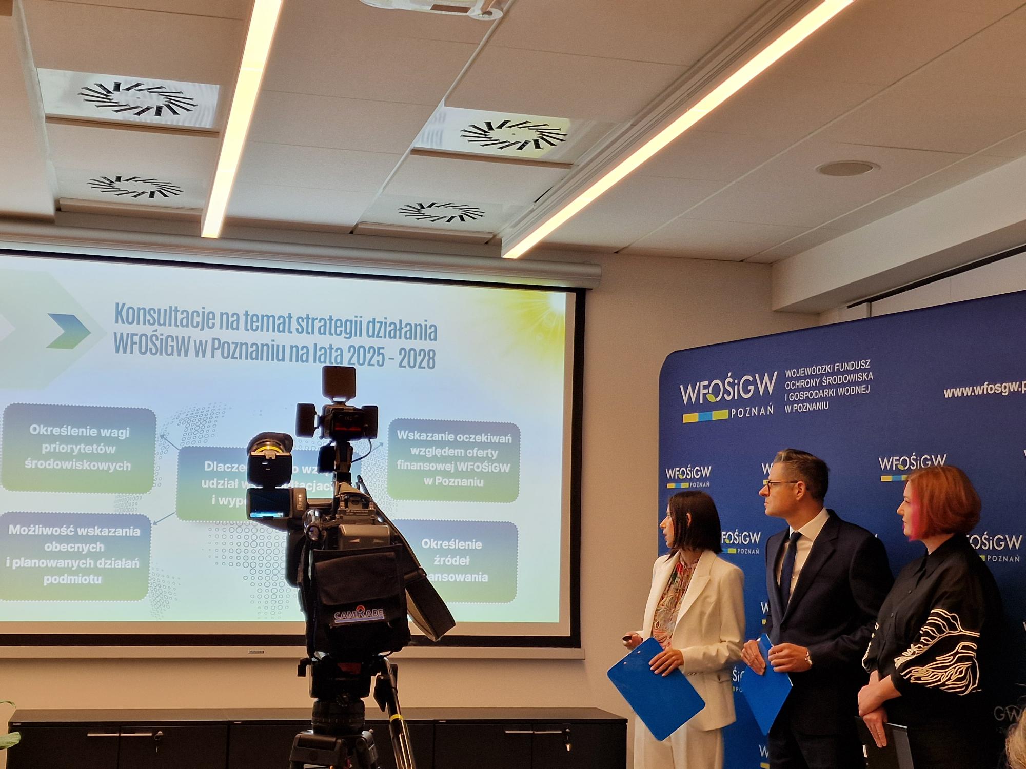 Zdjęcie przedstawia widok na wyświetlana prezentacja, a po prawej stronie, na tle ścianki z logotypami WFOŚiGW stoją 3 osoby i przemawiają. Na pierwszym planie widoczna jest kamera.