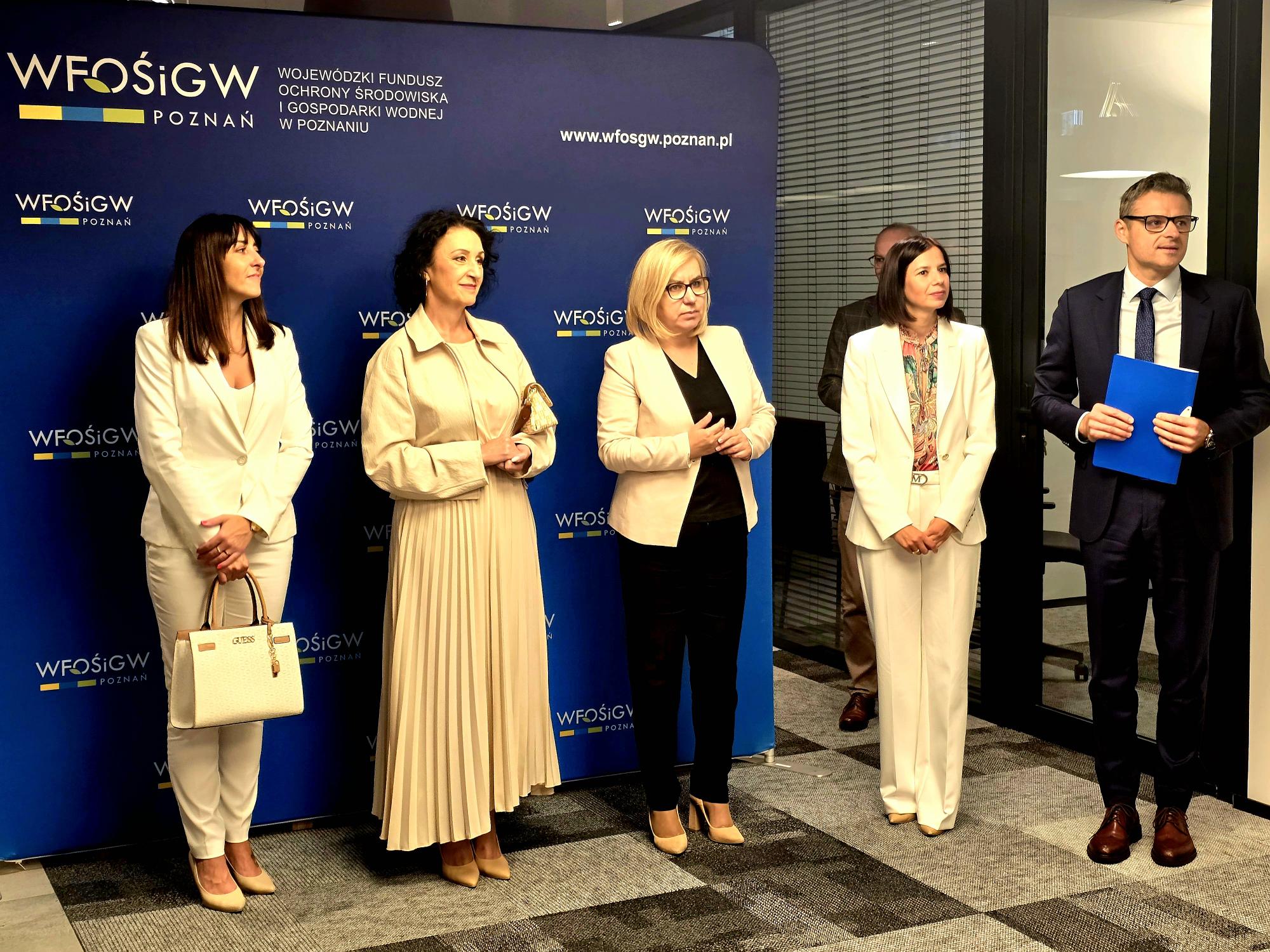 Na tle ścianki stoi 5 osób ,jdna osoba przemawia do zgromadzonych ludzi.