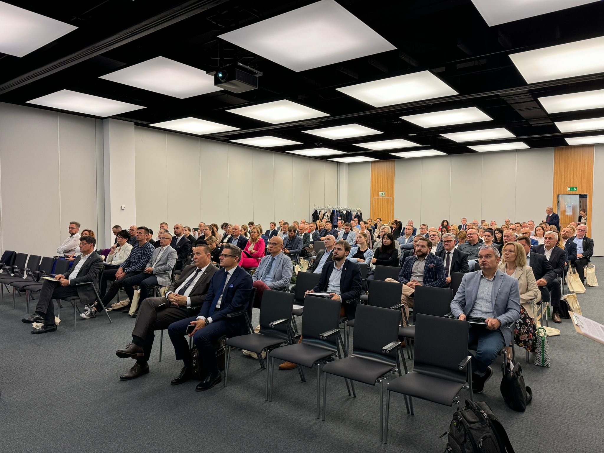 Widok na sale na konferencji, na której na krzesłach siedzą uczestnicy.
