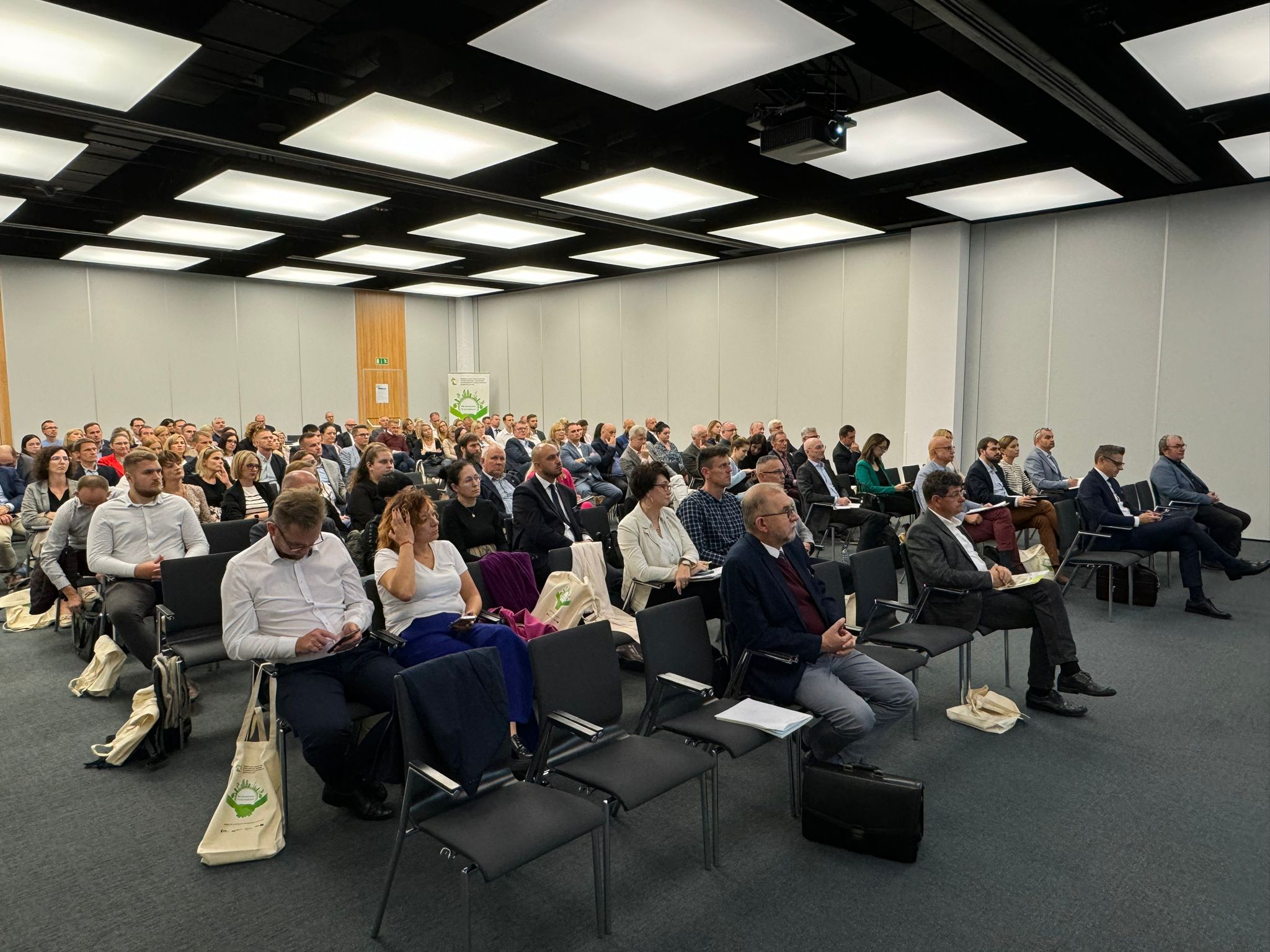 Widok na sale na konferencji, na której na krzesłach siedzą uczestnicy.