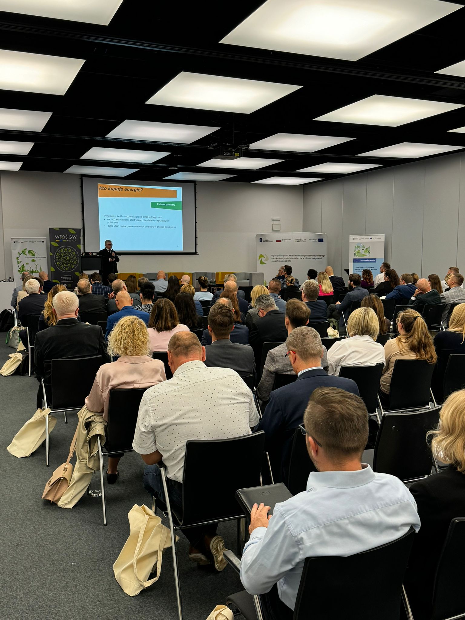 Widok na sale na konferencji, na której na krzesłach siedzą uczestnicy.