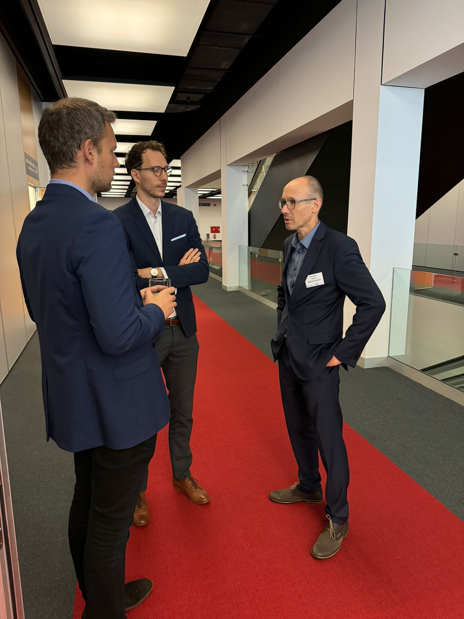 3 osoby stoją razem w hallu przed salą konferencyjną i rozmawiają.