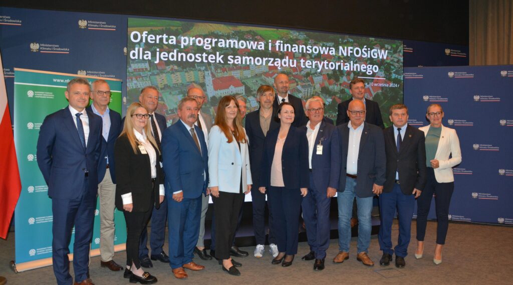 Na zdjęciu na tle dużego ekranu z nazwą spotkania stoi grupa uczestników spotkania.