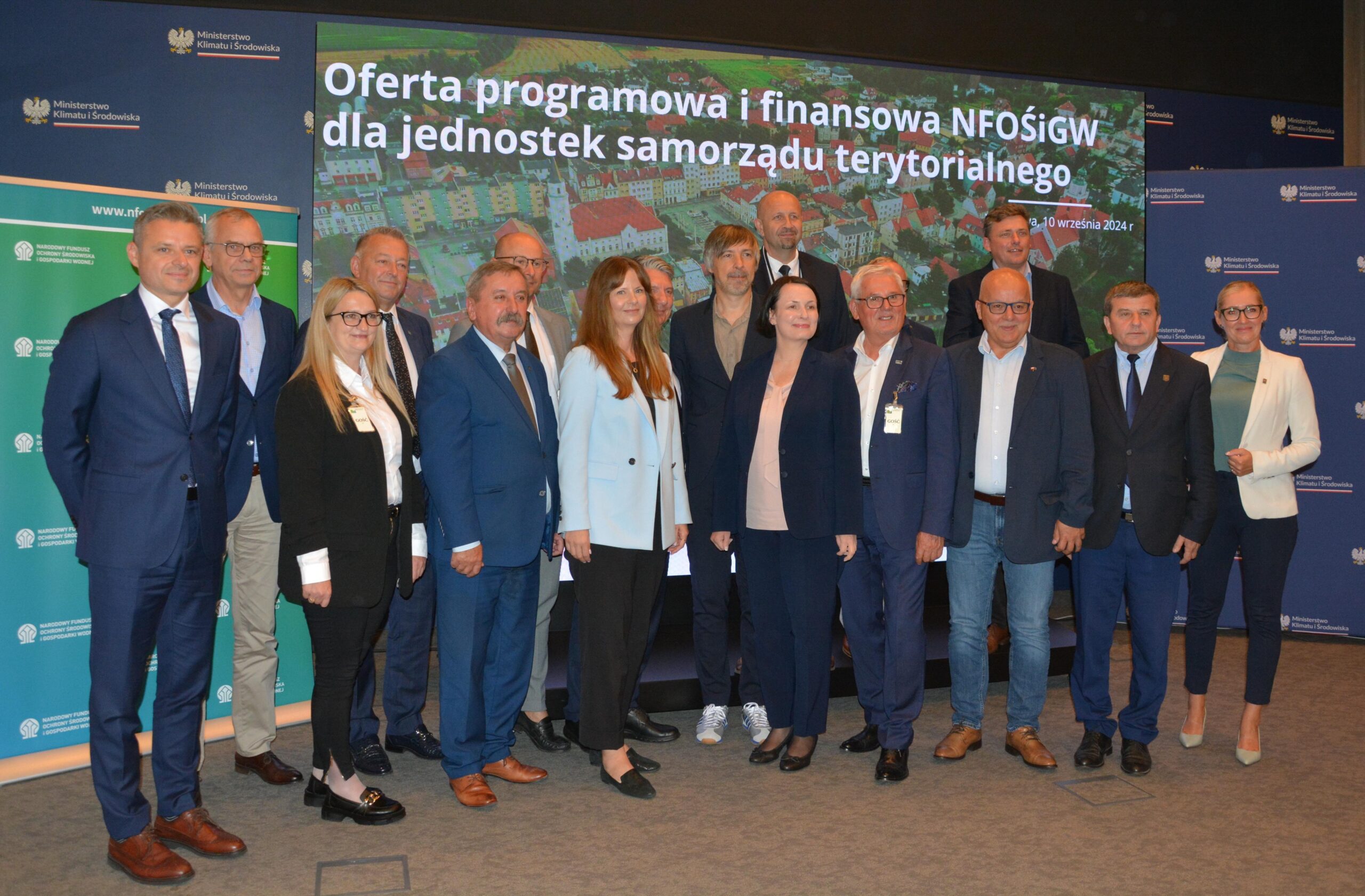 Na zdjęciu na tle dużego ekranu z nazwą spotkania stoi grupa uczestników spotkania.